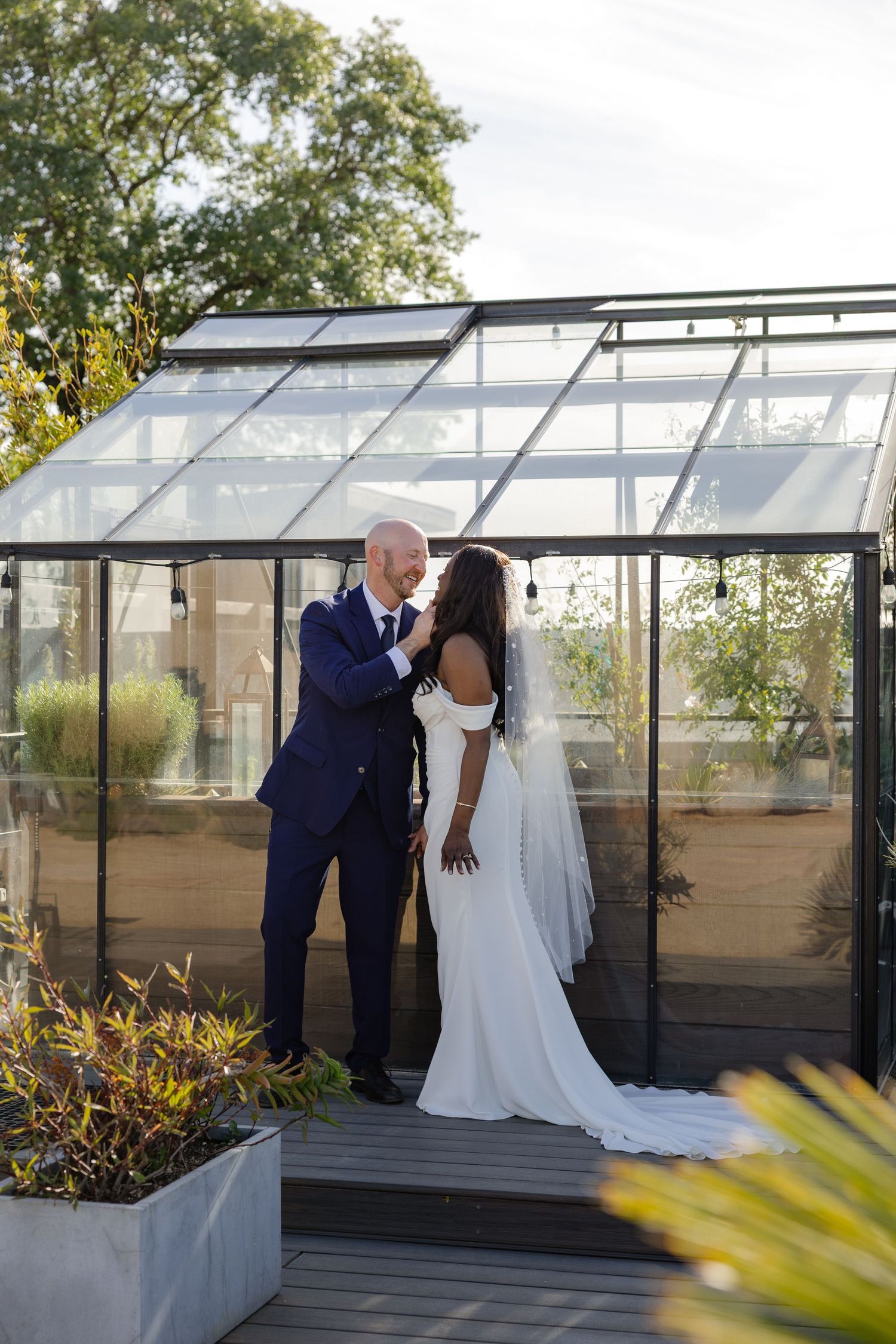 Bride and groom share a romantic moment in front of a glass greenhouse on a rooftop deck on their wedding day.