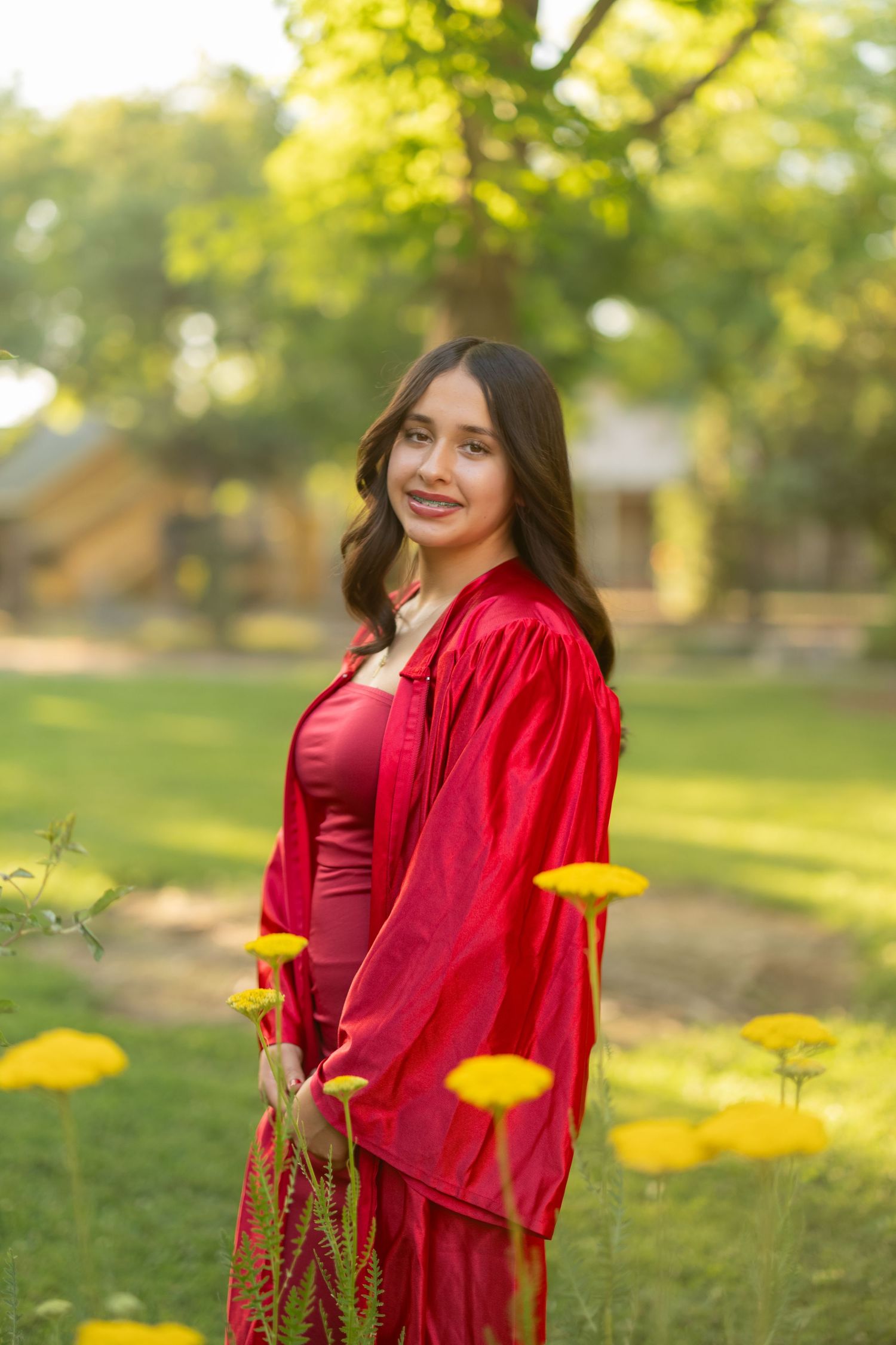 Graduate in red attire stands among yellow flowers in sunlit garden setting.