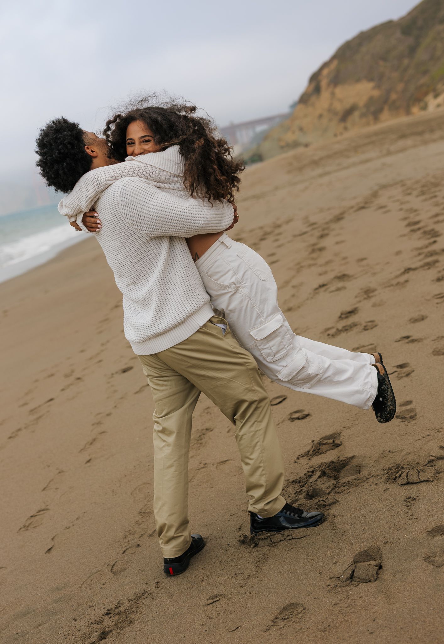 A couple embraces in a joyful hug on a cloudy beach with cliffs in the background.
