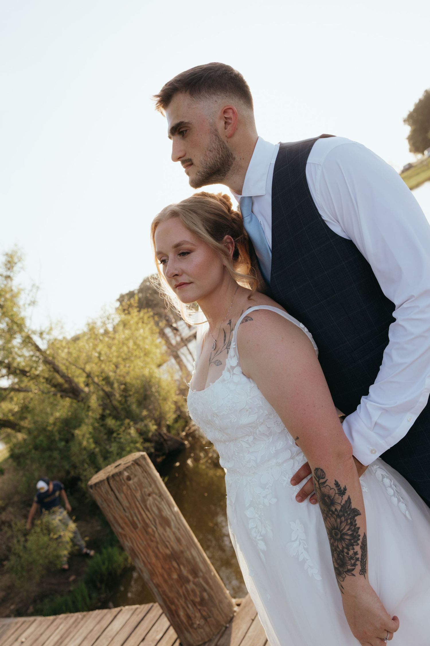 A bride and groom pose romantically on a wooden deck outdoors, the groom embracing the bride from behind in wedding attire.