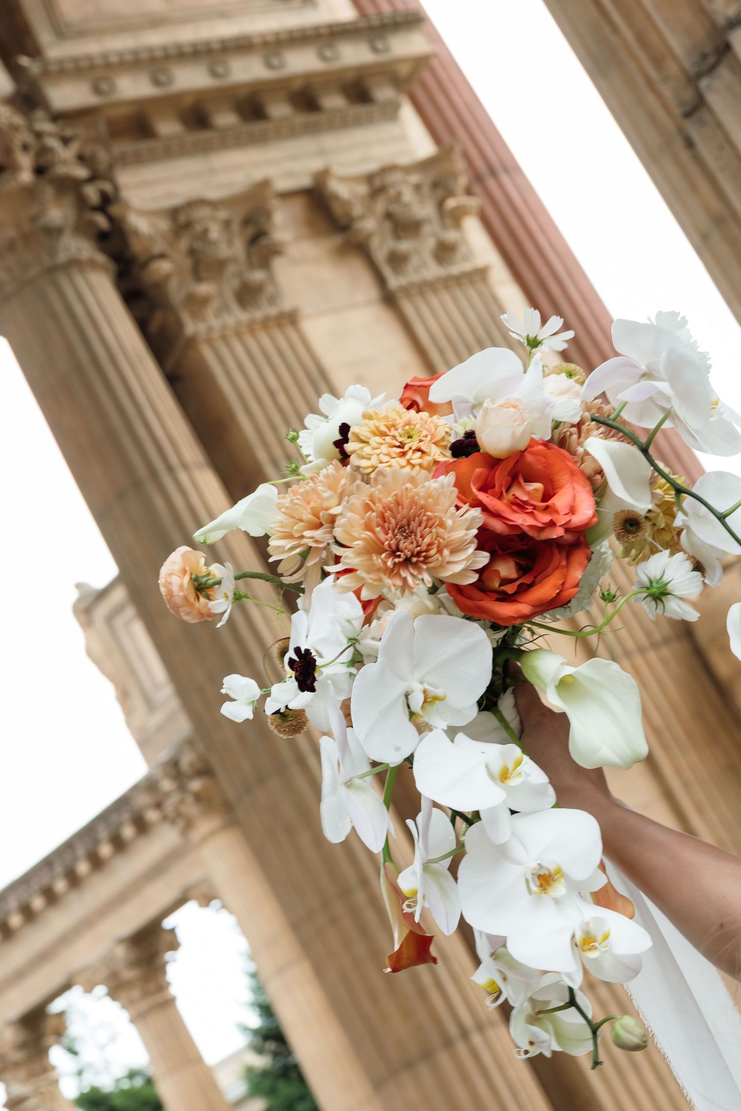 Bridal bouquet with white orchids, orange roses, and peach dahlias held before classical stone columns.
