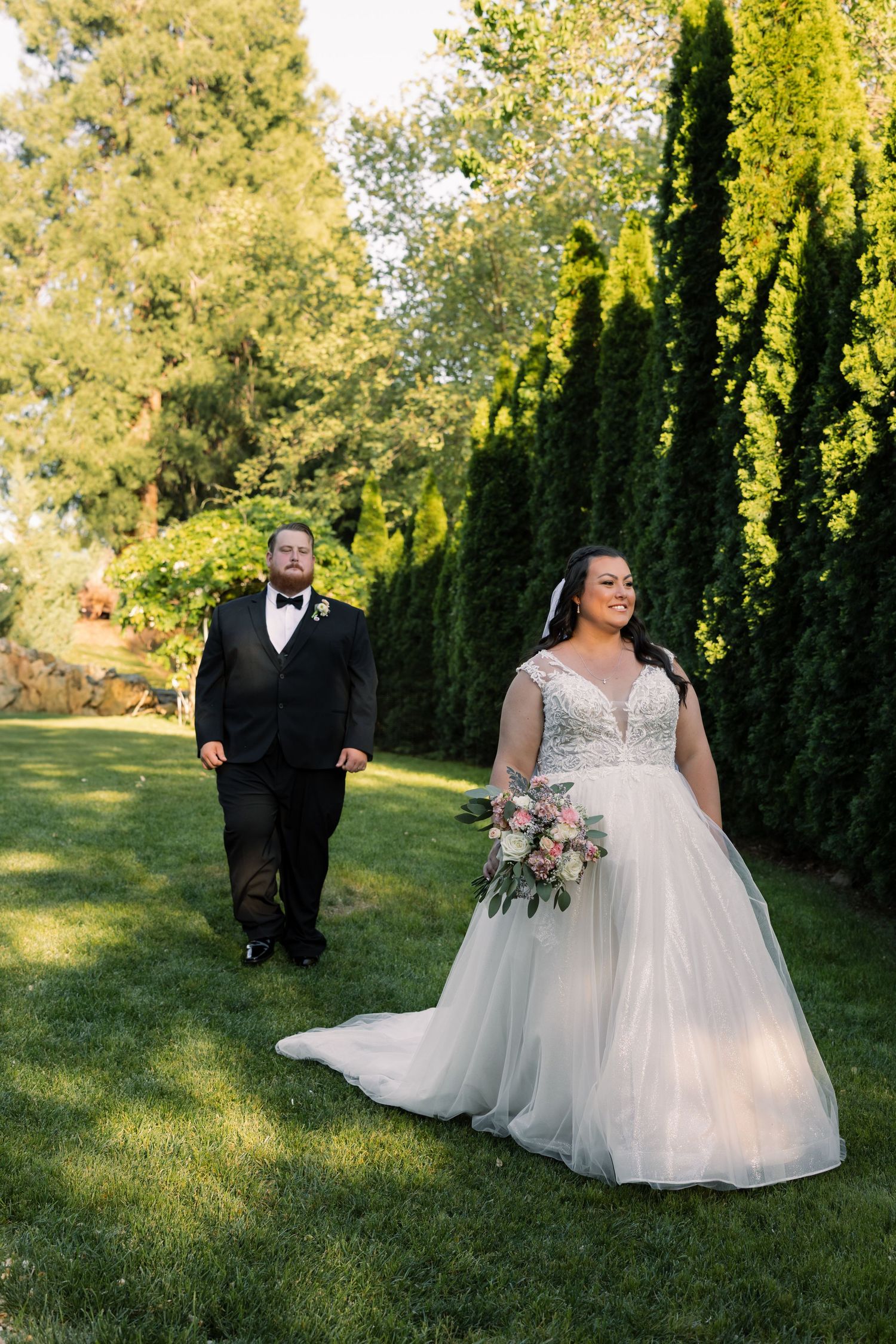 A couple poses for a wedding photo on a lawn with tall cypress trees in the background.