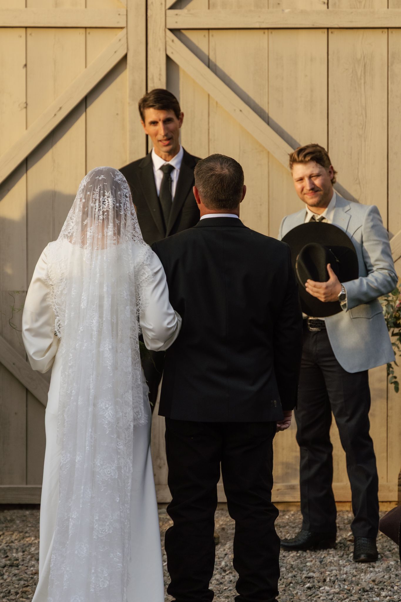 A wedding party lines up against a wooden fence backdrop with the bride in a flowing white veil and groomsmen in dark suits.