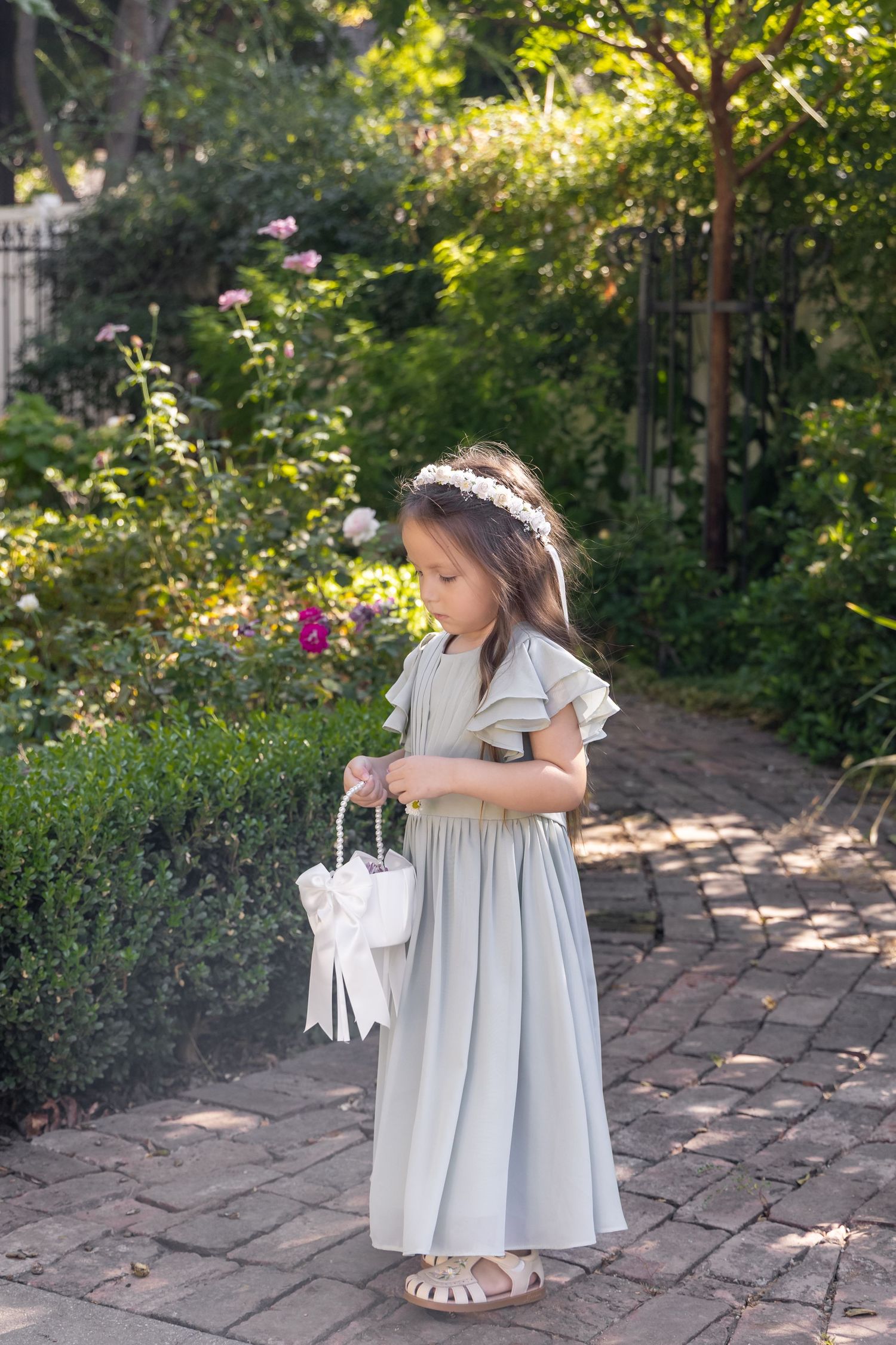 Young flower girl in light gray dress twirls on brick pathway in garden.
