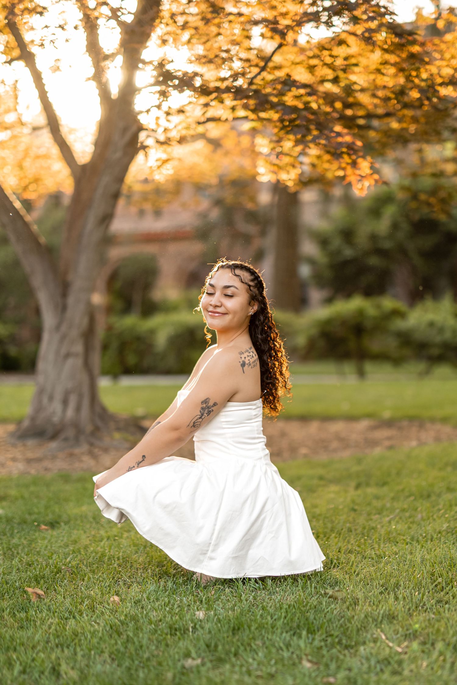 A person in a white dress twirls gracefully on green grass during golden hour as sunlight filters through trees.