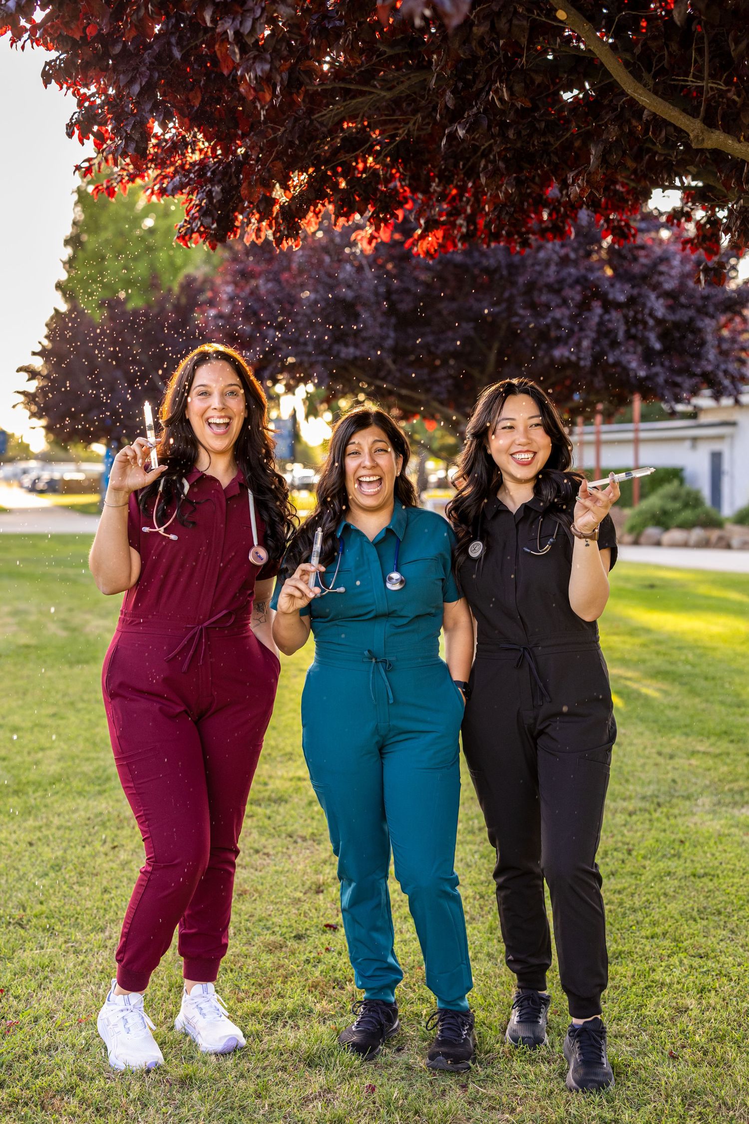 Three healthcare workers in colored scrubs jump and celebrate outdoors.