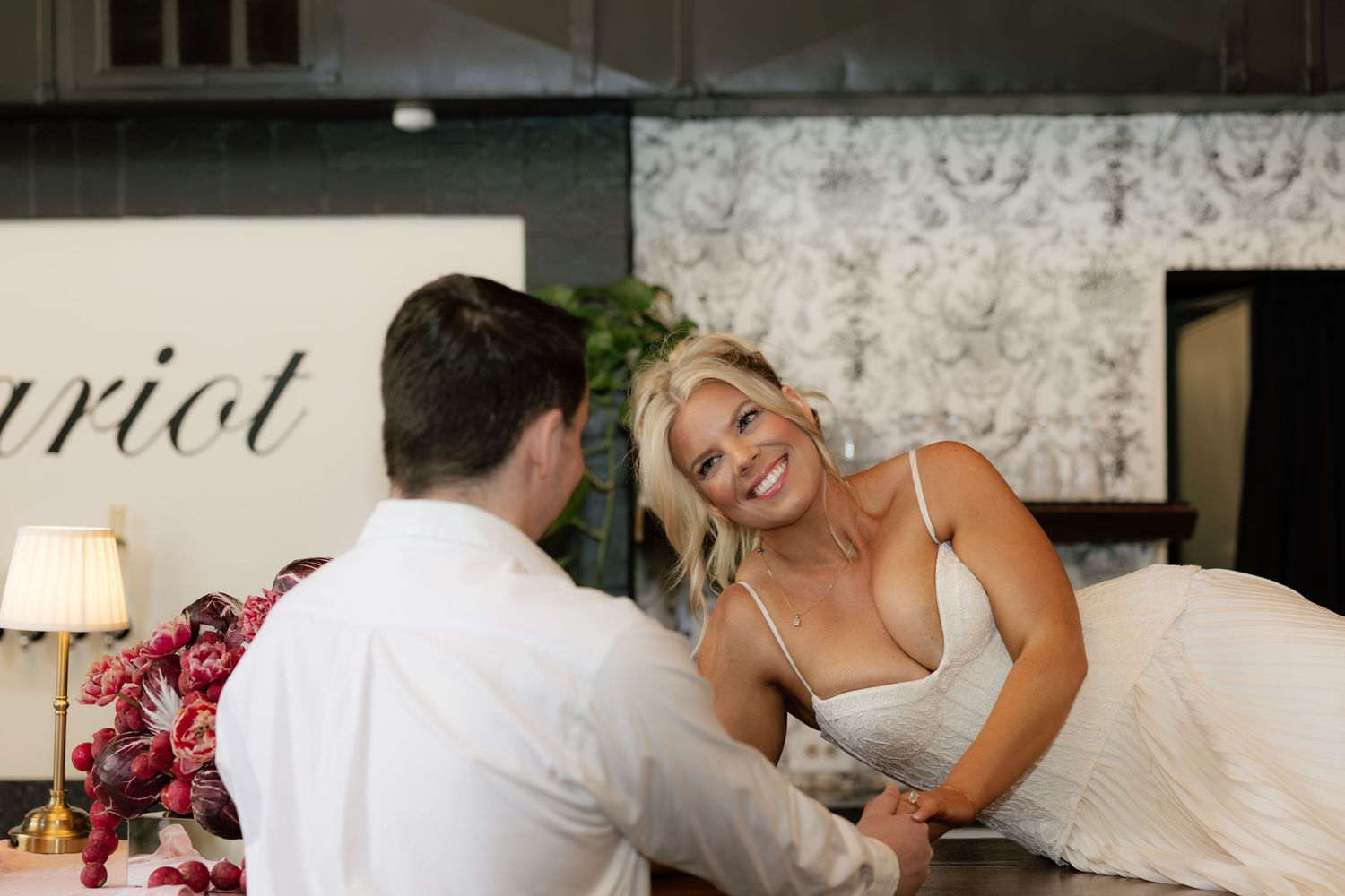 Smiling bride in white gown holds hands with groom at wedding reception table with floral decor.