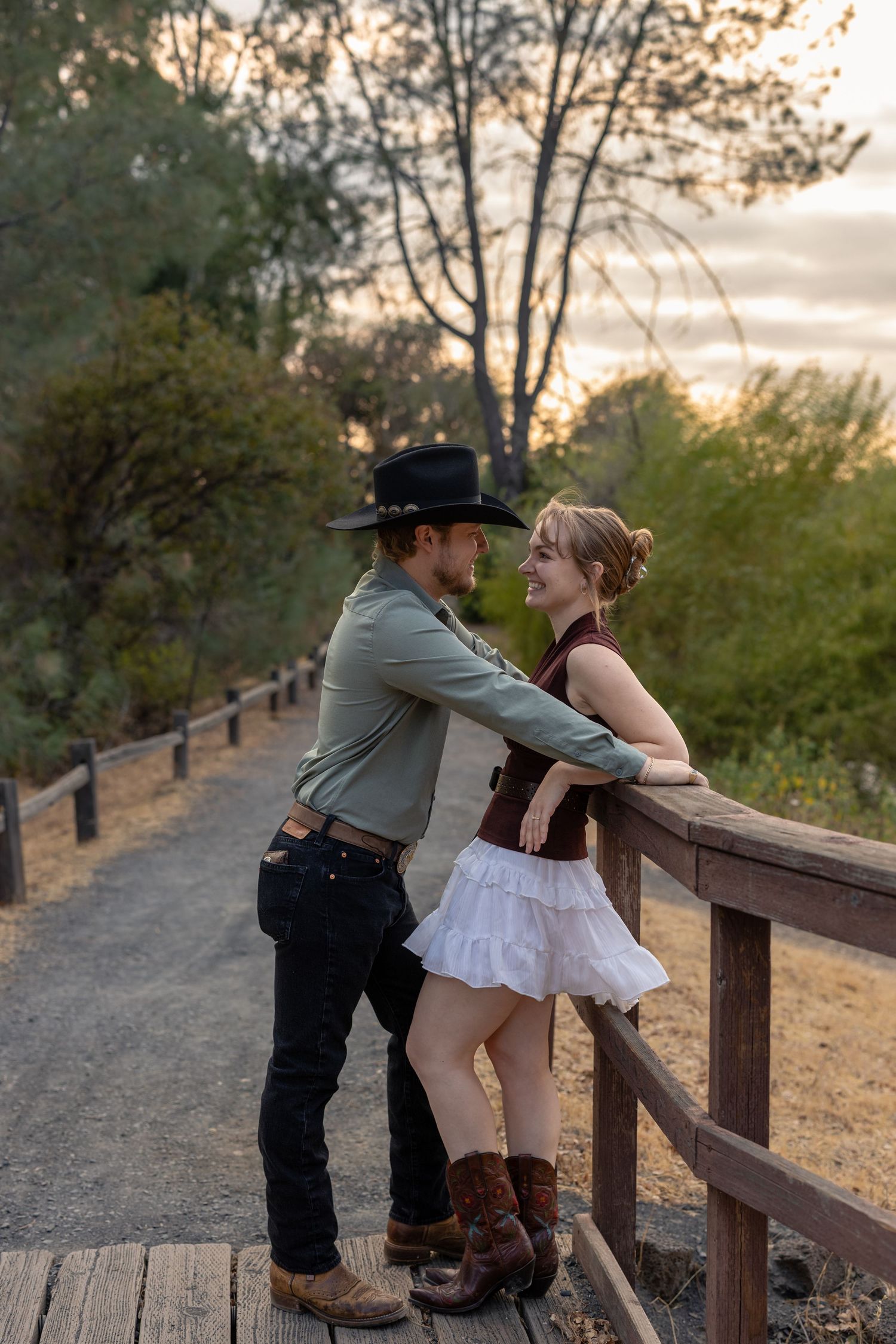 A playful couple in western attire shares intimate moments along a wooden fence at sunset.