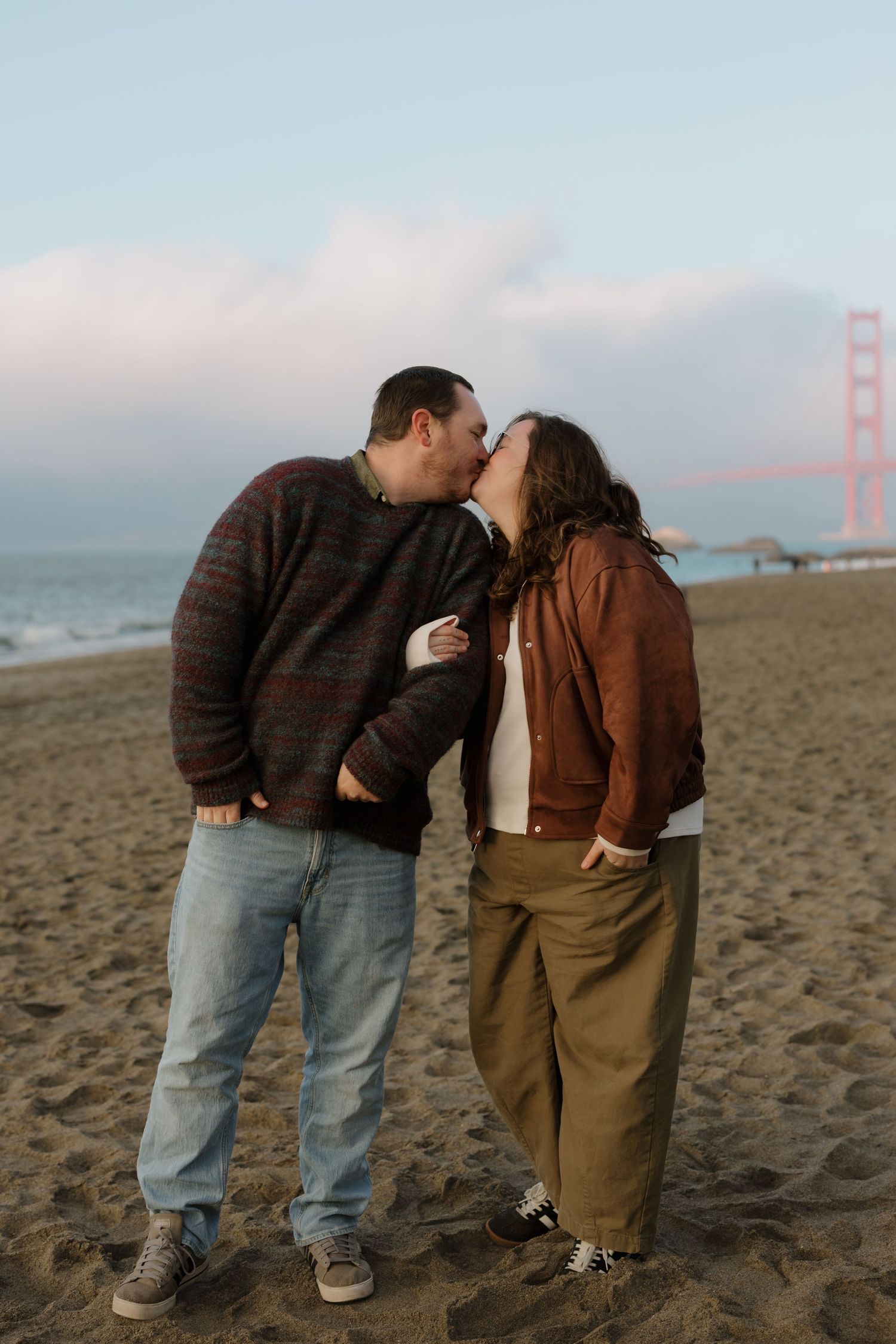 A couple shares an intimate moment on a beach with Golden Gate Bridge visible in the distance.