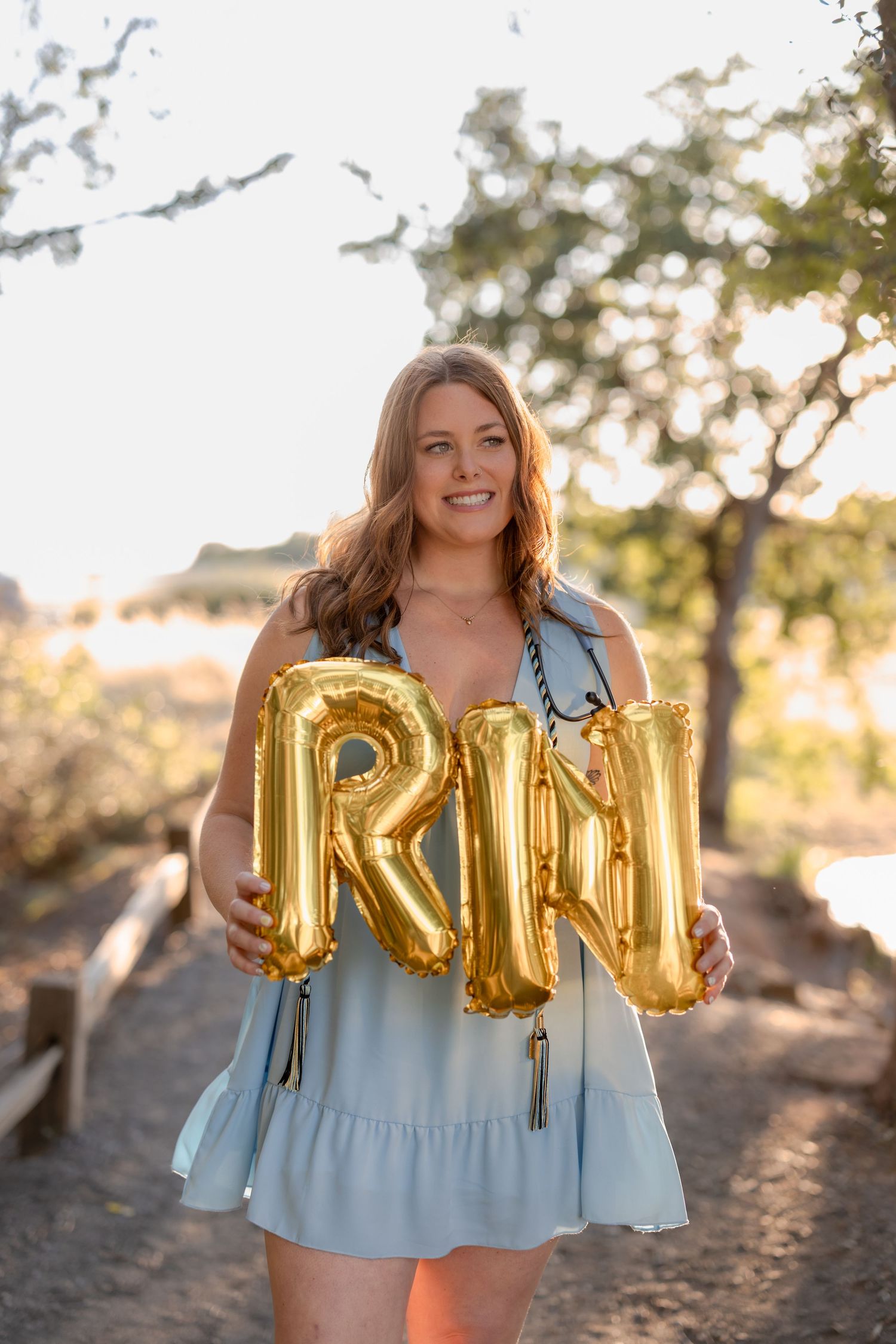 Person holds gold RN balloon letters while wearing light blue dress outdoors.