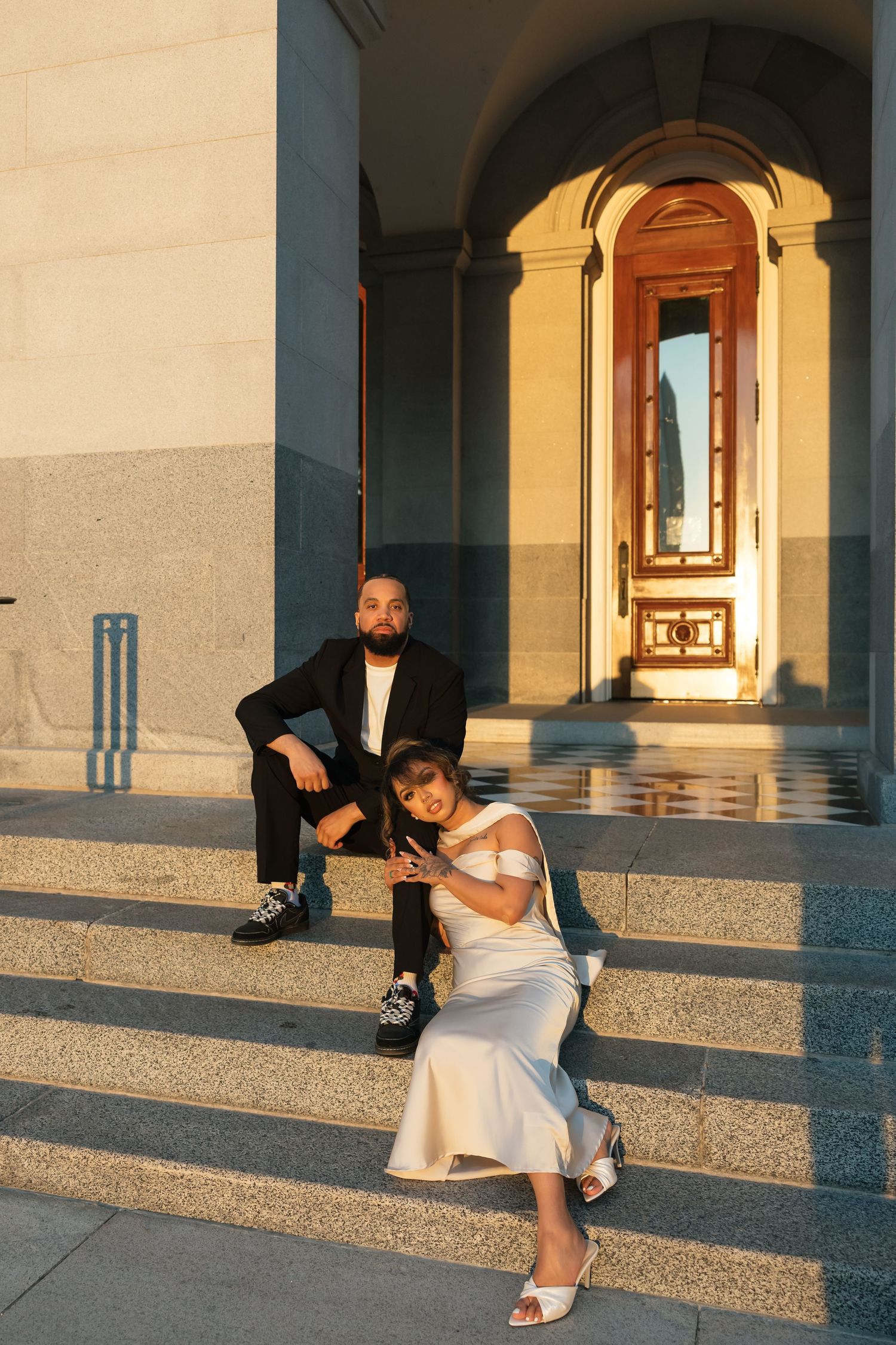 Bride in white dress and groom in black suit pose on stone steps of elegant building at golden hour.