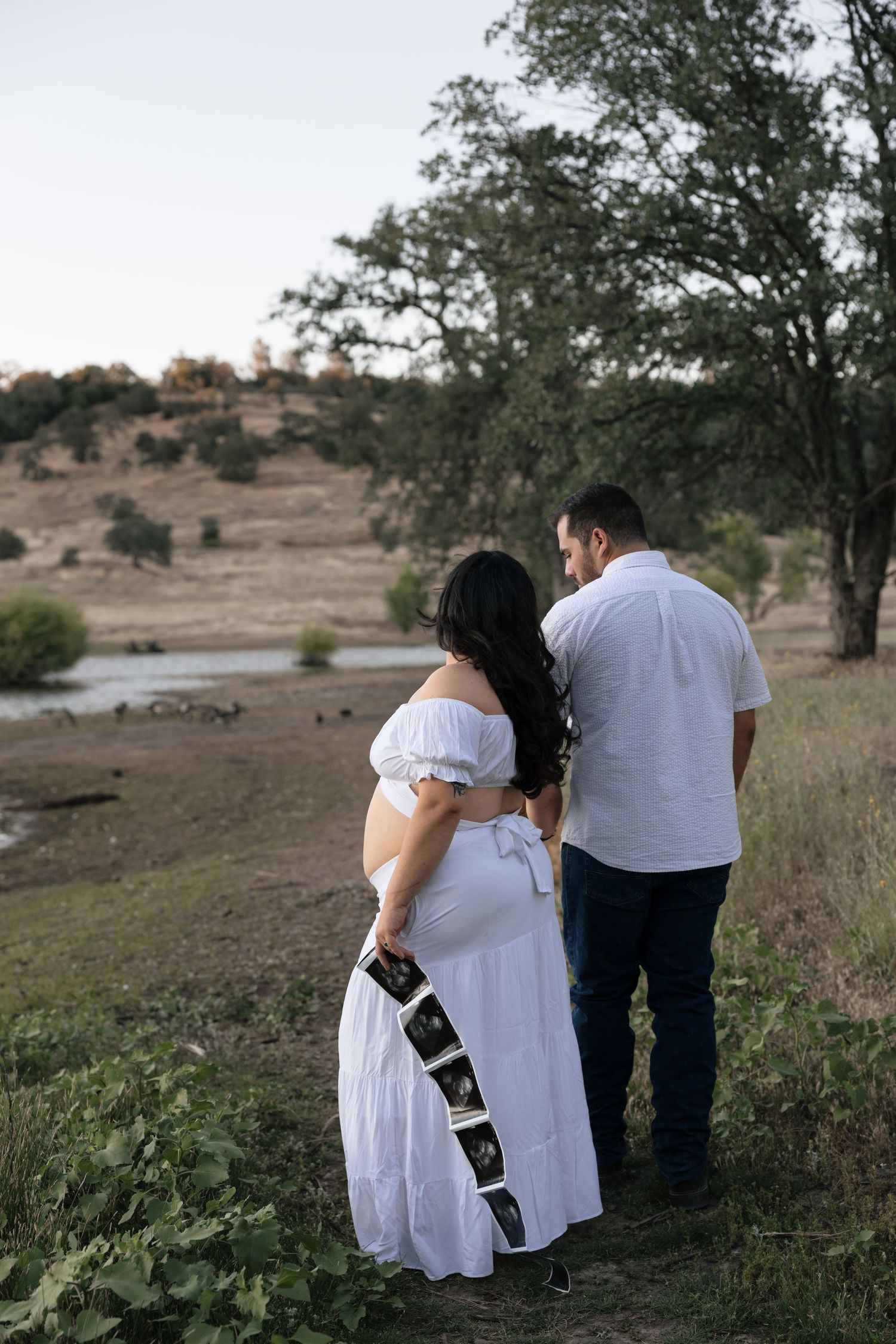 Pregnant couple holding ultrasound photos outdoors near a lake, woman in white off-shoulder dress, maternity photography.