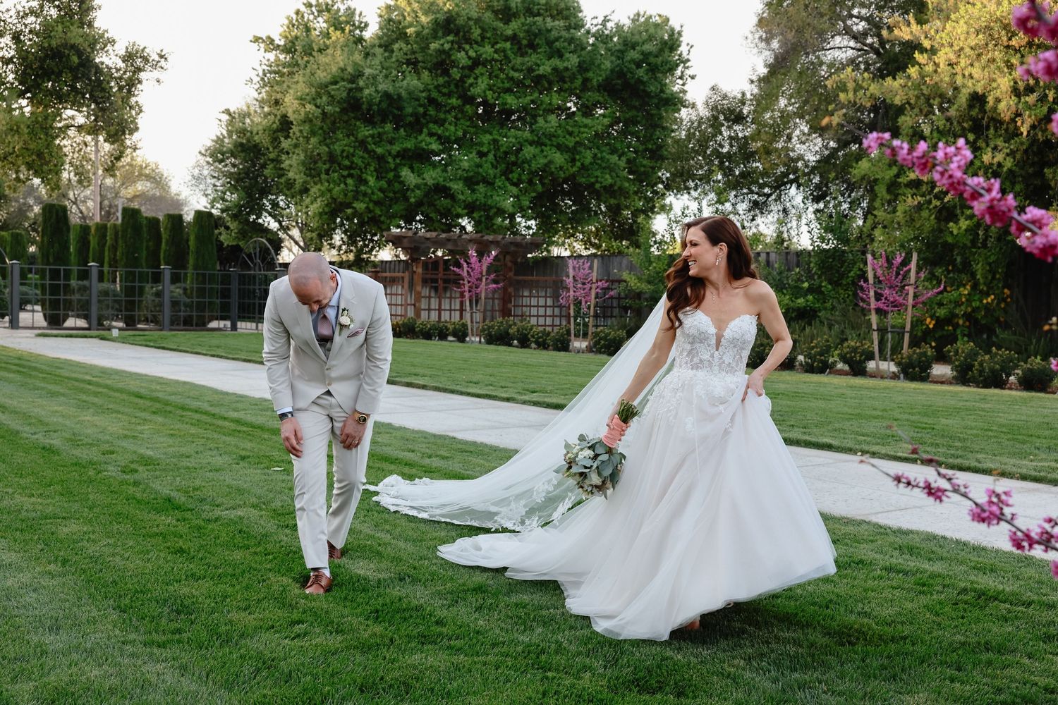 A couple in wedding attire walks on a manicured lawn surrounded by blooming flowers and trees in an outdoor garden setting.