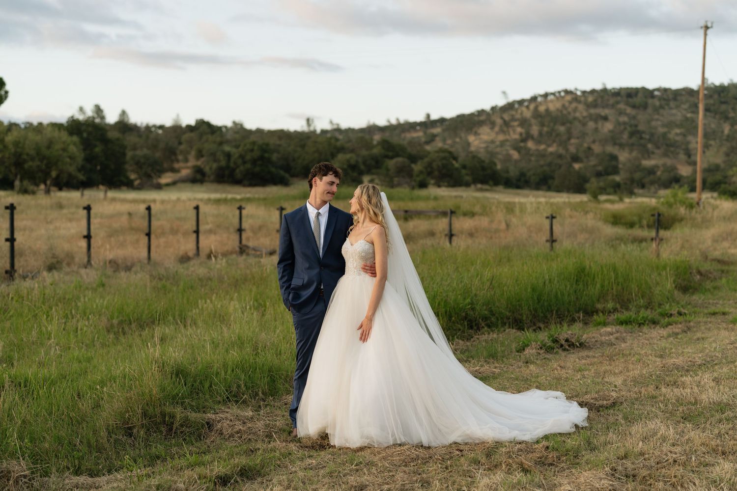 Bride and groom stand together in a lush green field with rolling hills at golden hour during outdoor wedding.