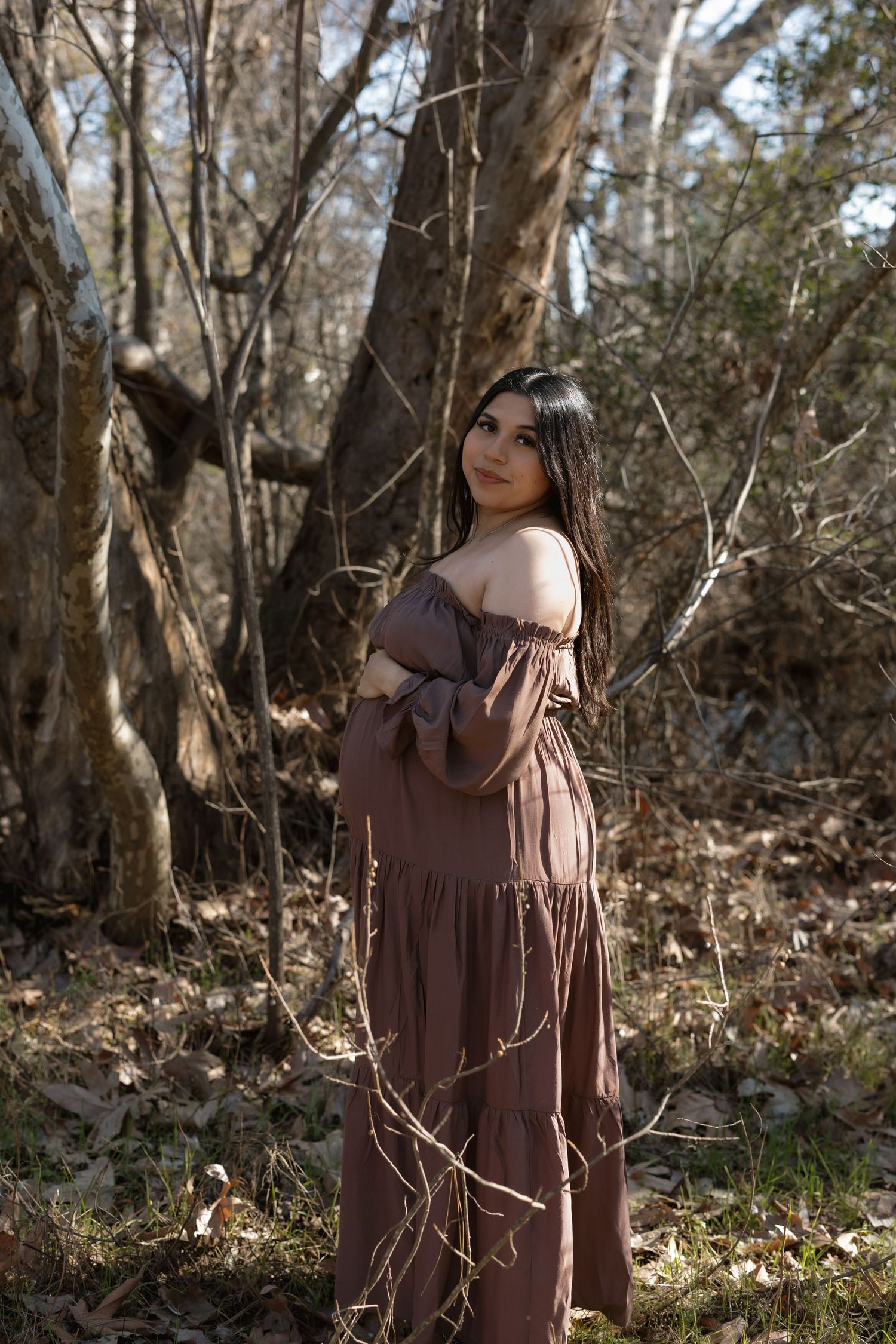 A person in a brown dress poses thoughtfully among bare trees in a forest setting during autumn.