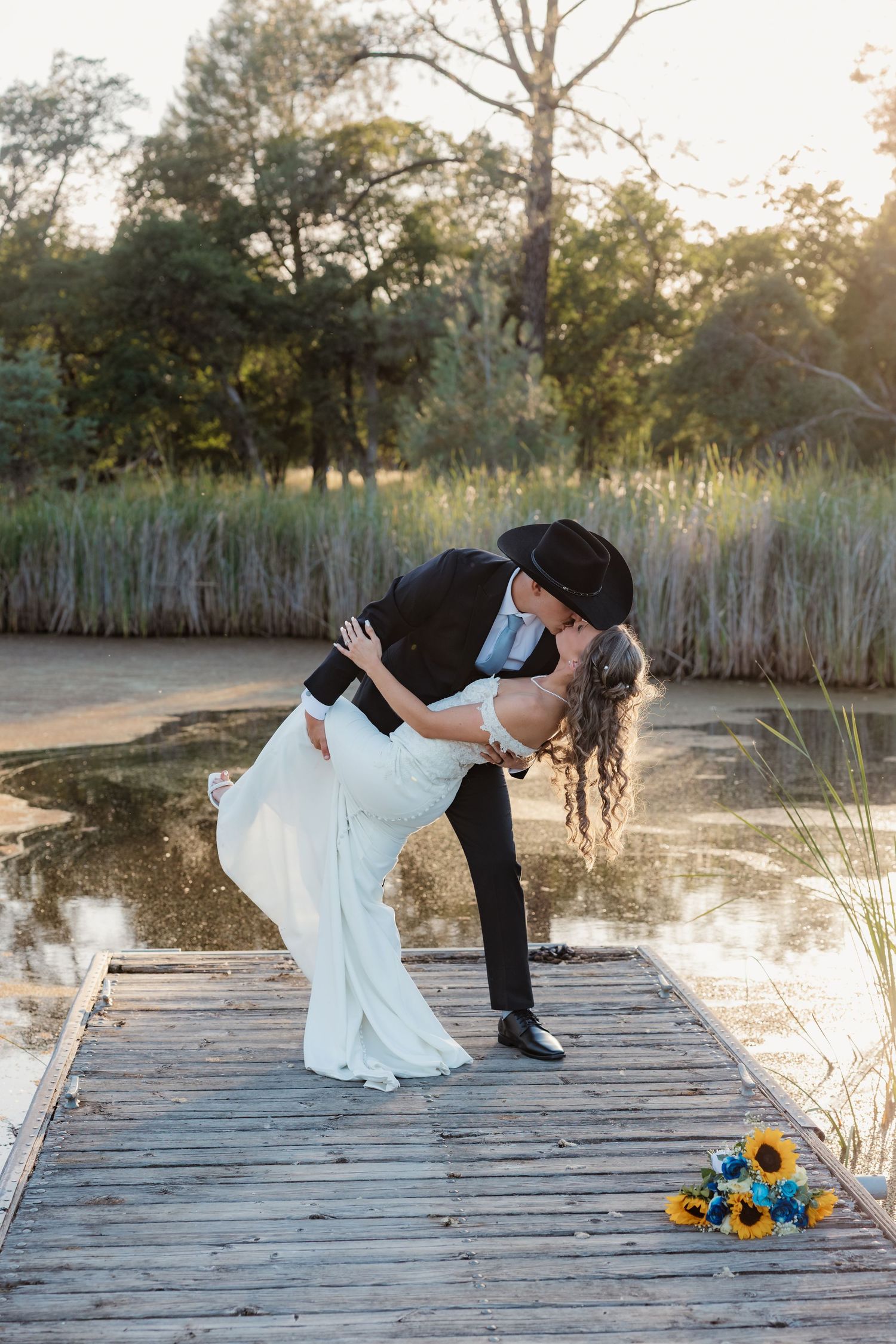 Groom in black cowboy hat dips bride in white gown for a romantic kiss on a wooden dock by a serene pond.