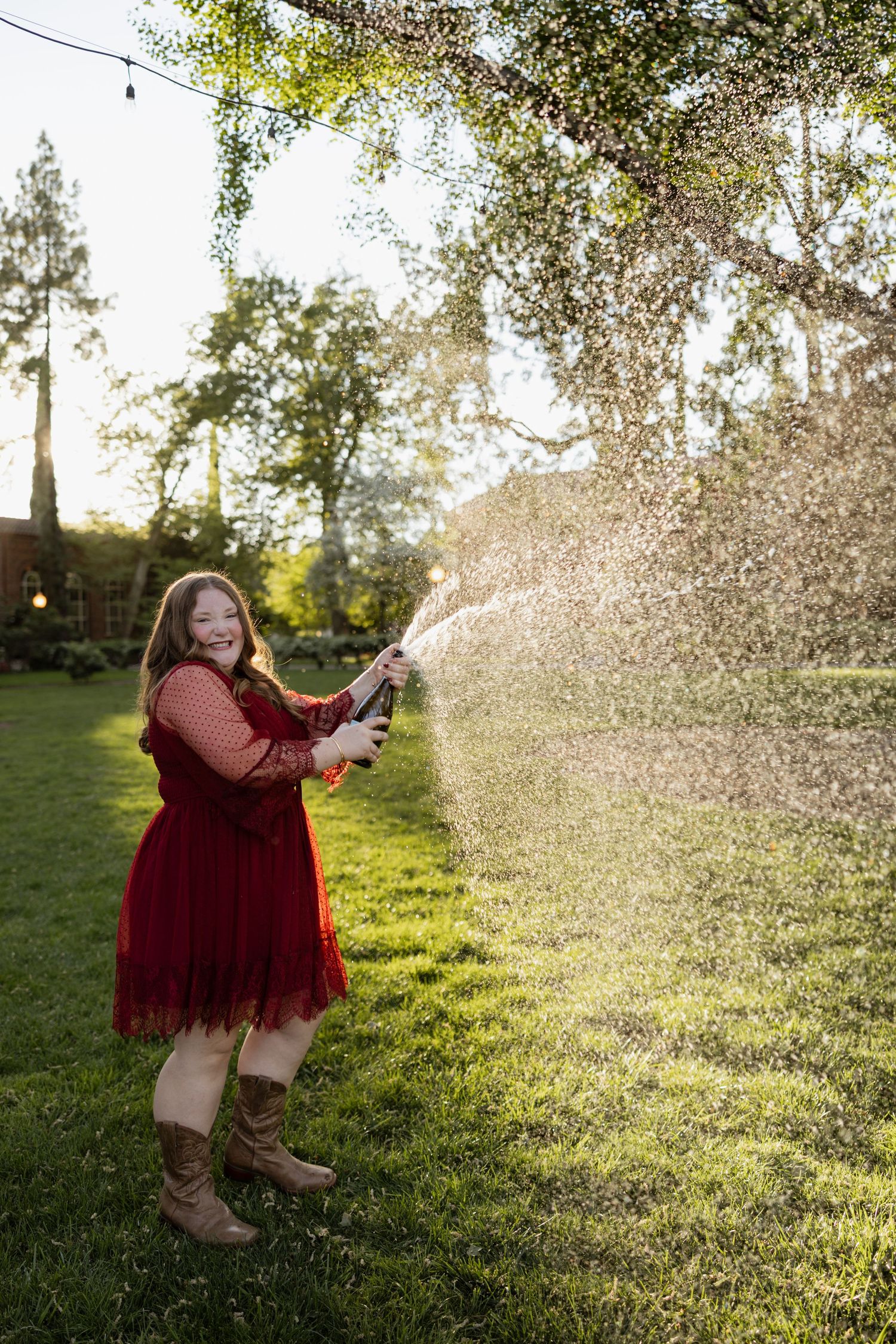 Young woman in red dress and boots joyfully spraying water from garden hose in sunny backyard.