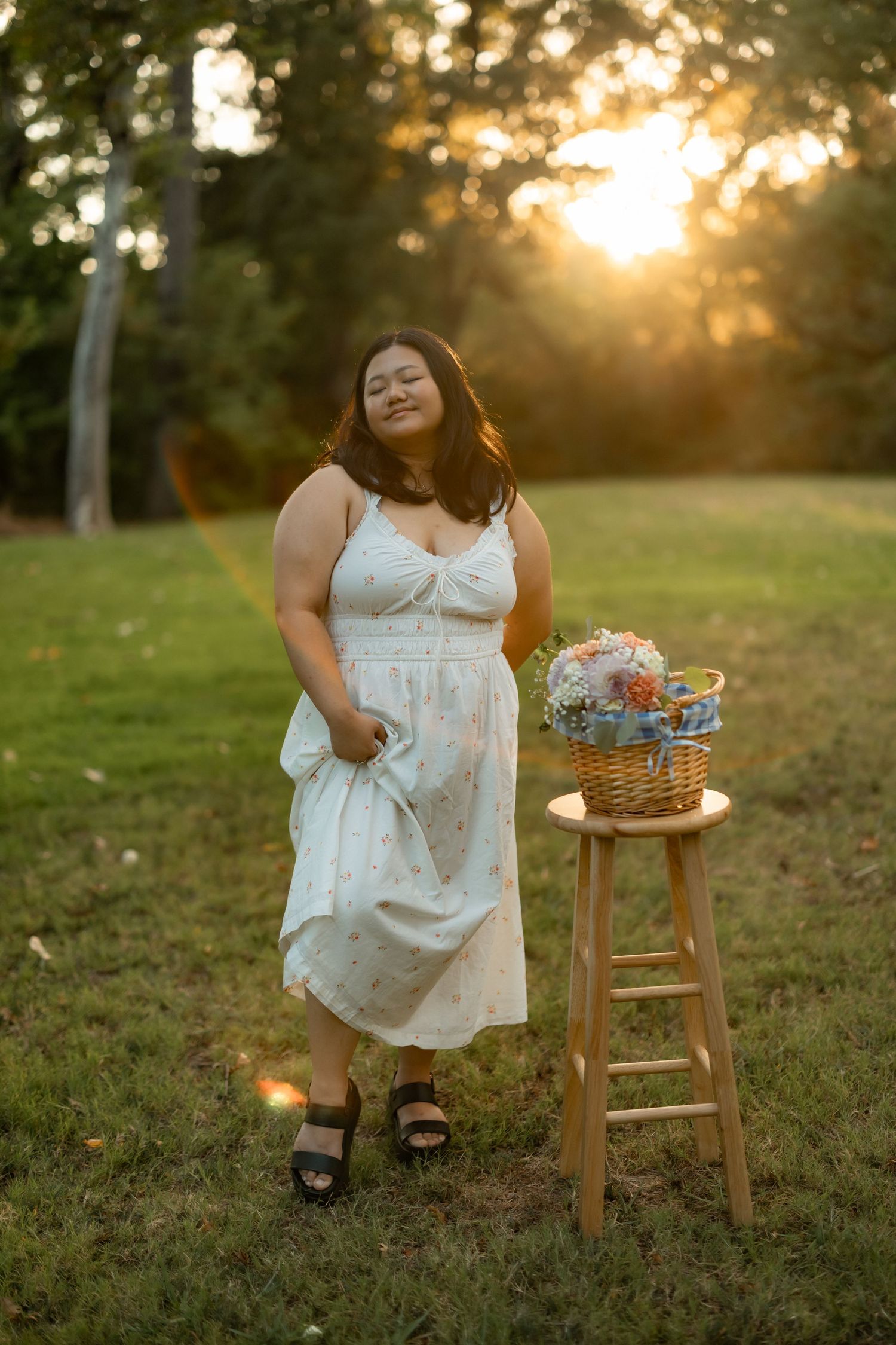 Woman in white dress poses with vintage wooden stool in sunlit field.