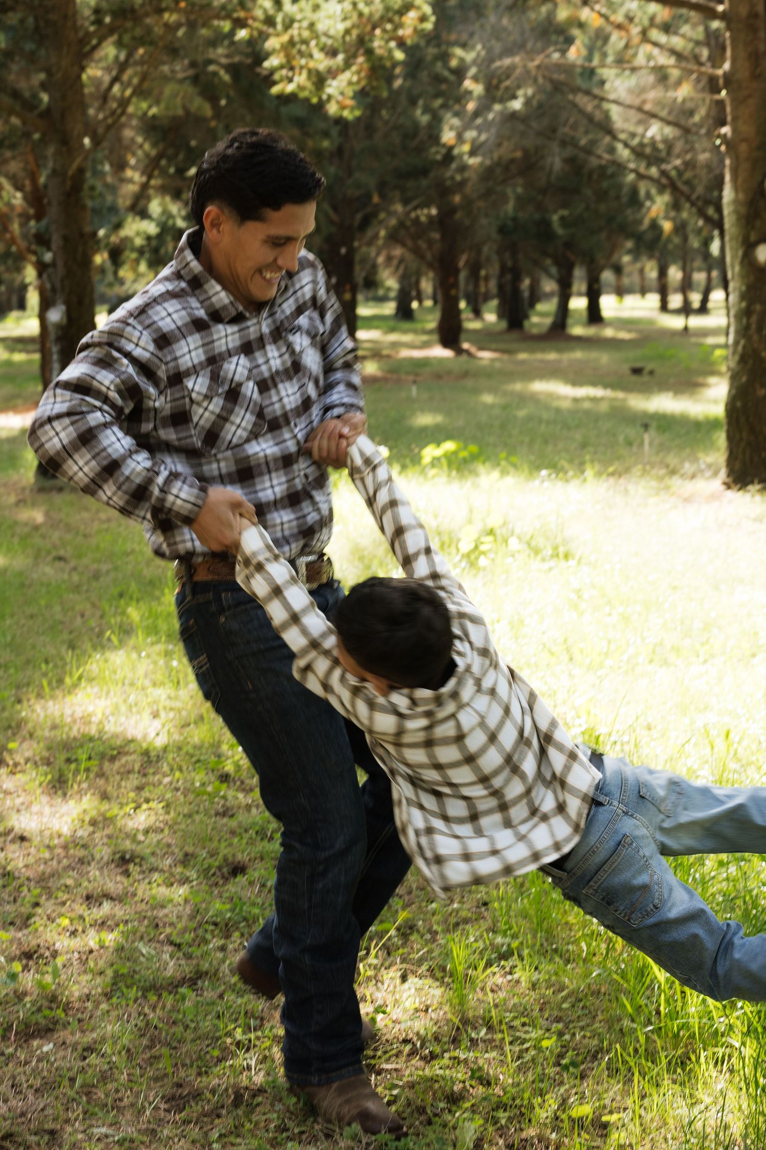 A person in a plaid shirt swings a child playfully in a sunny park setting with trees in the background.
