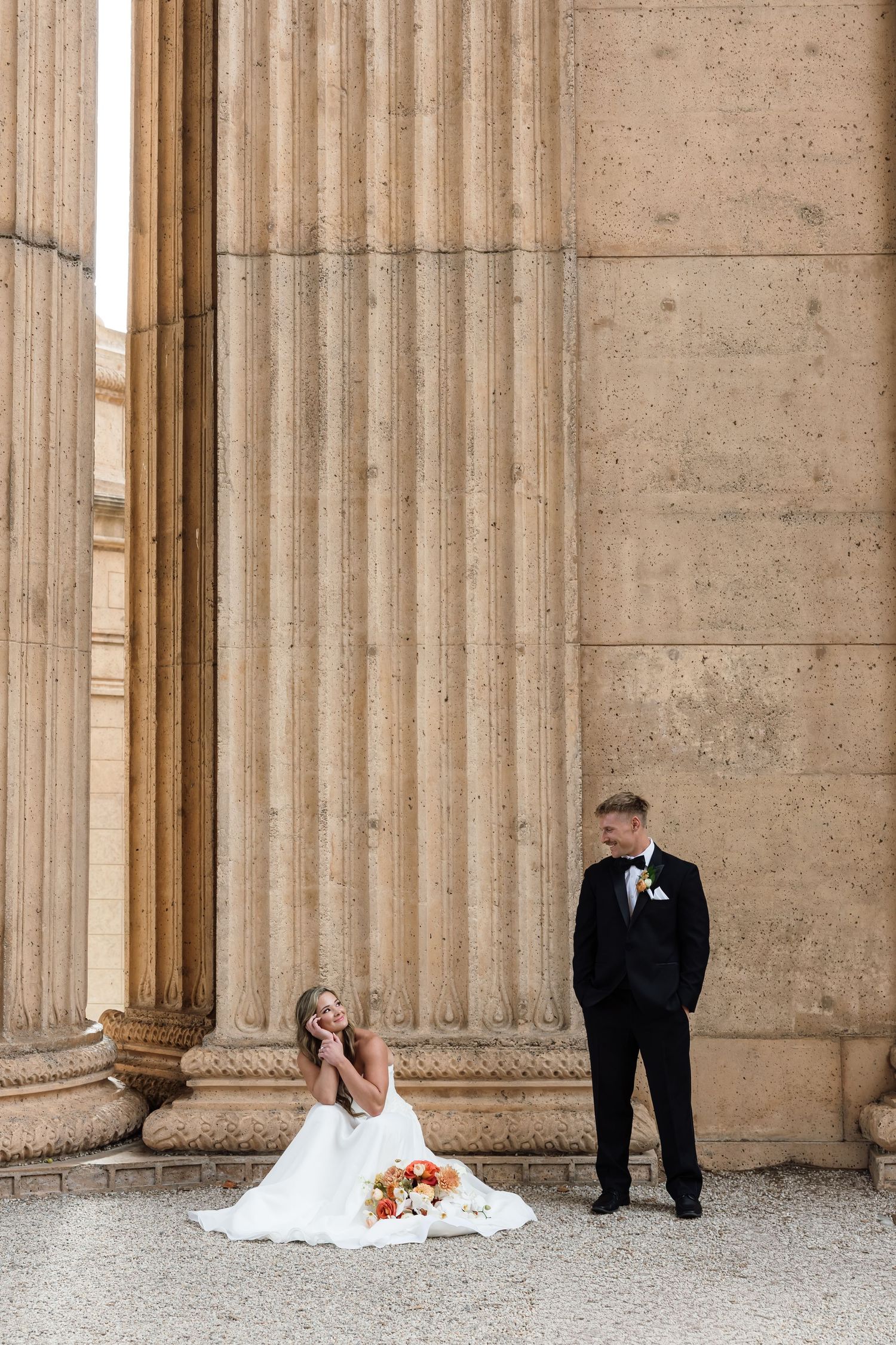Bride in white gown sits on ground beside groom in tuxedo against grand stone columns at Palace of Fine Arts.