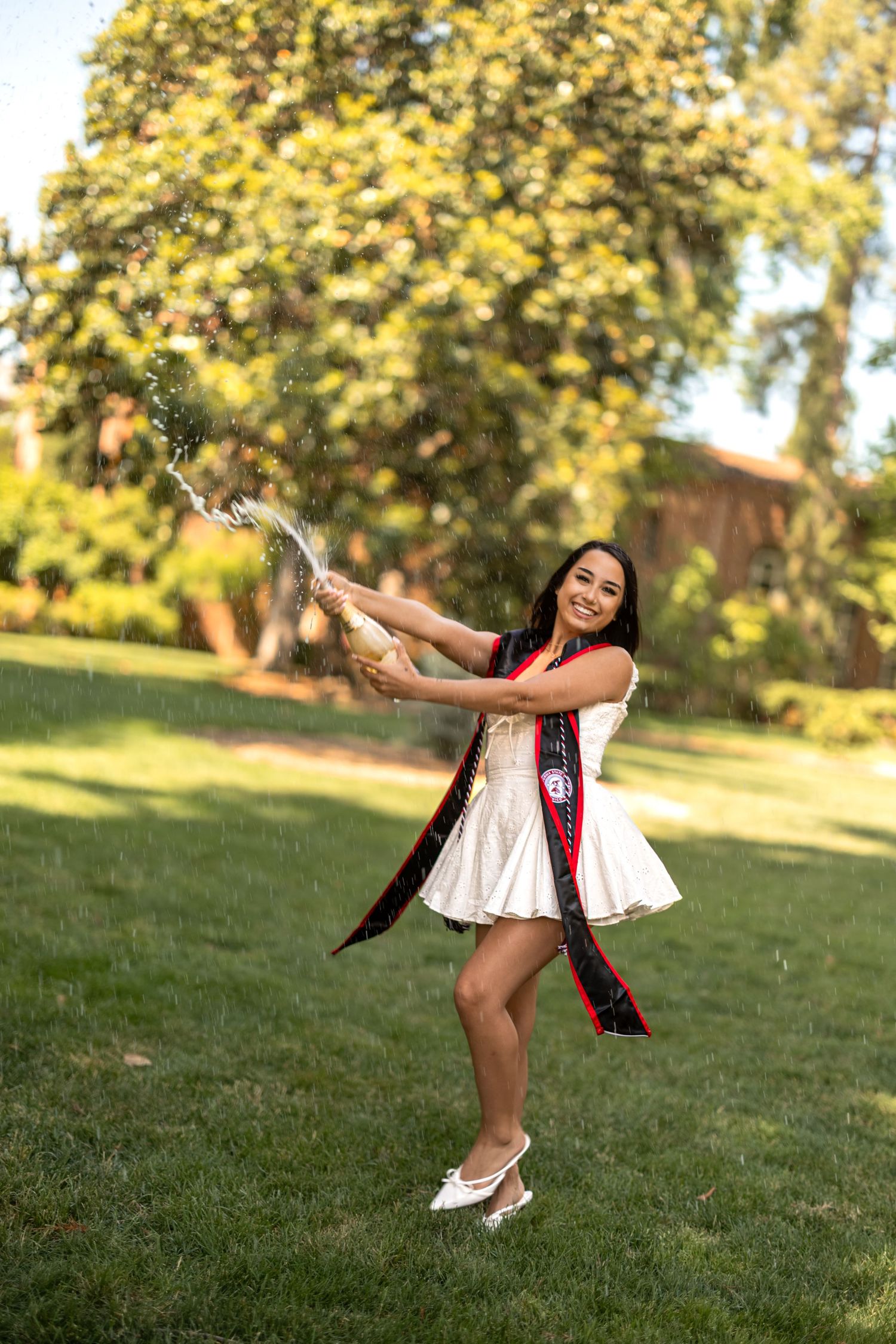 Graduate in white dress and graduation stole celebrates on sunny lawn with joyful pose.