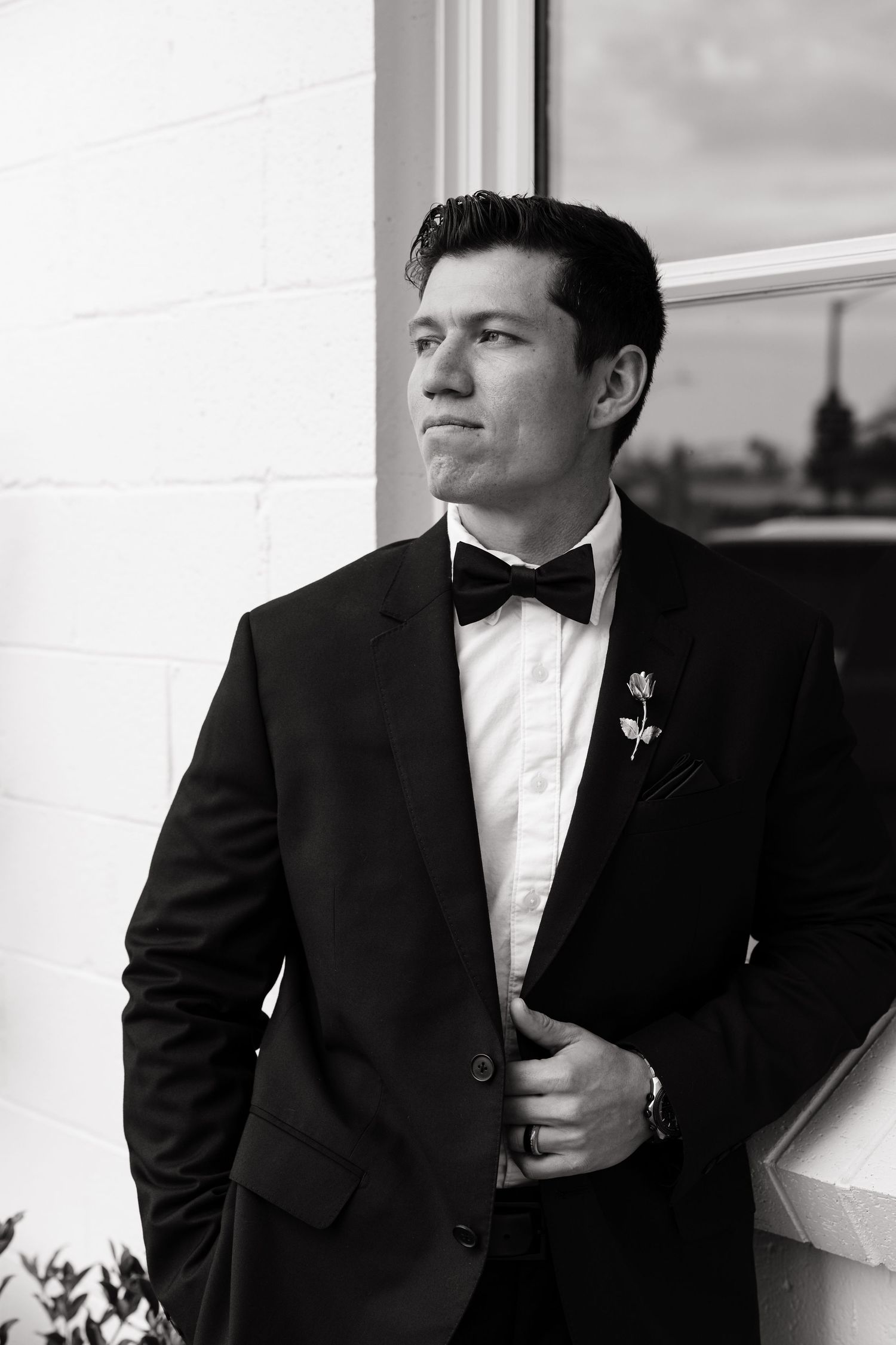 Black and white photo of a person in formal attire with bow tie standing against a light colored wall.