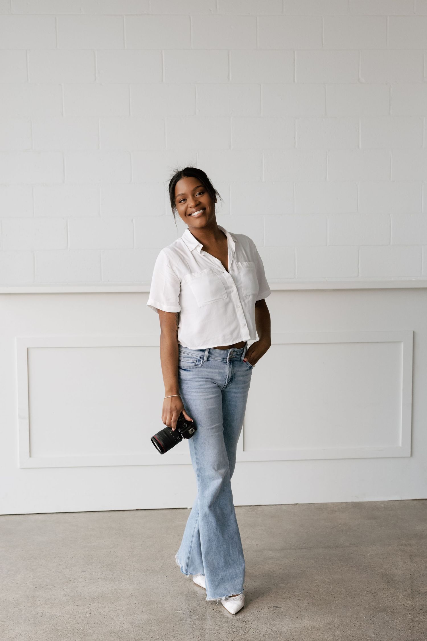 Person wearing white button-down shirt and light blue jeans poses against white wall in casual fashion photoshoot.
