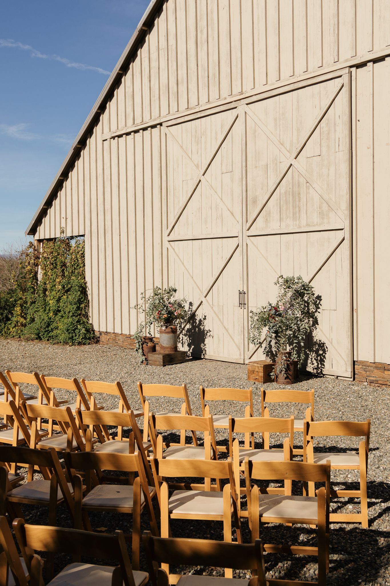 Wooden folding chairs lined up outside a rustic white barn for an outdoor event on a sunny day.