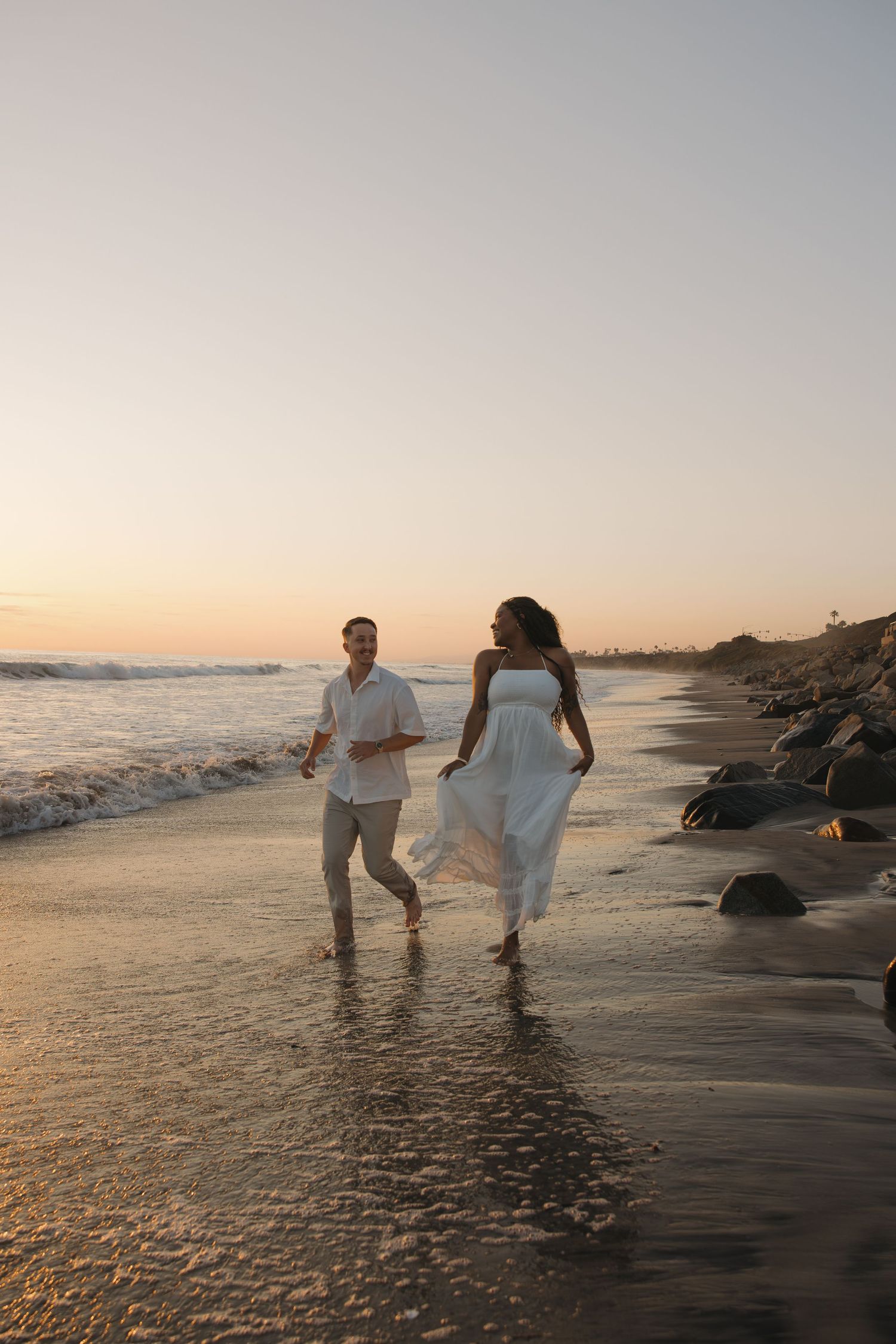 A couple walks along a beach at sunset with waves gently lapping at their feet, creating a romantic silhouette.