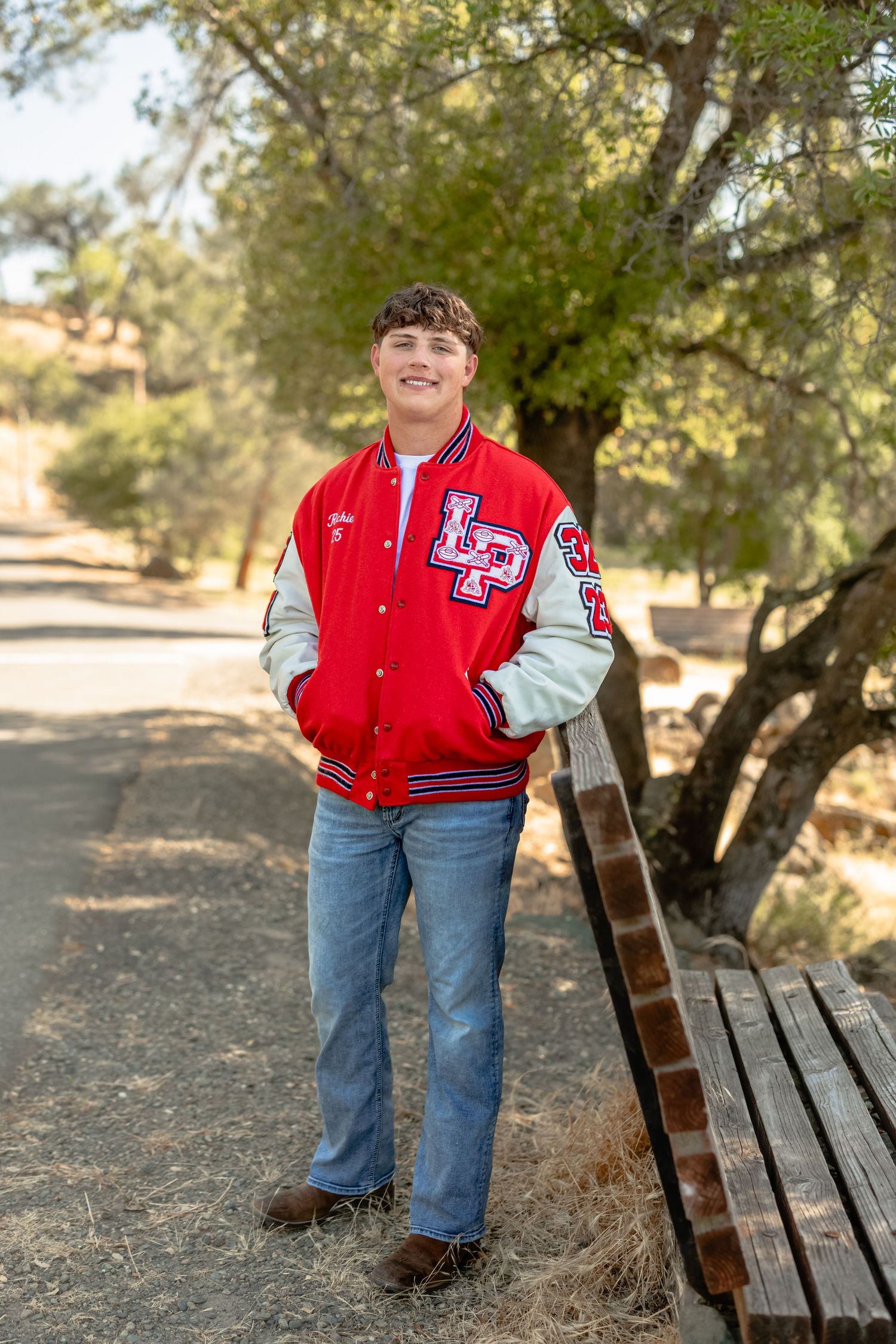 Student in red varsity letterman jacket poses on trail with wooden bench.