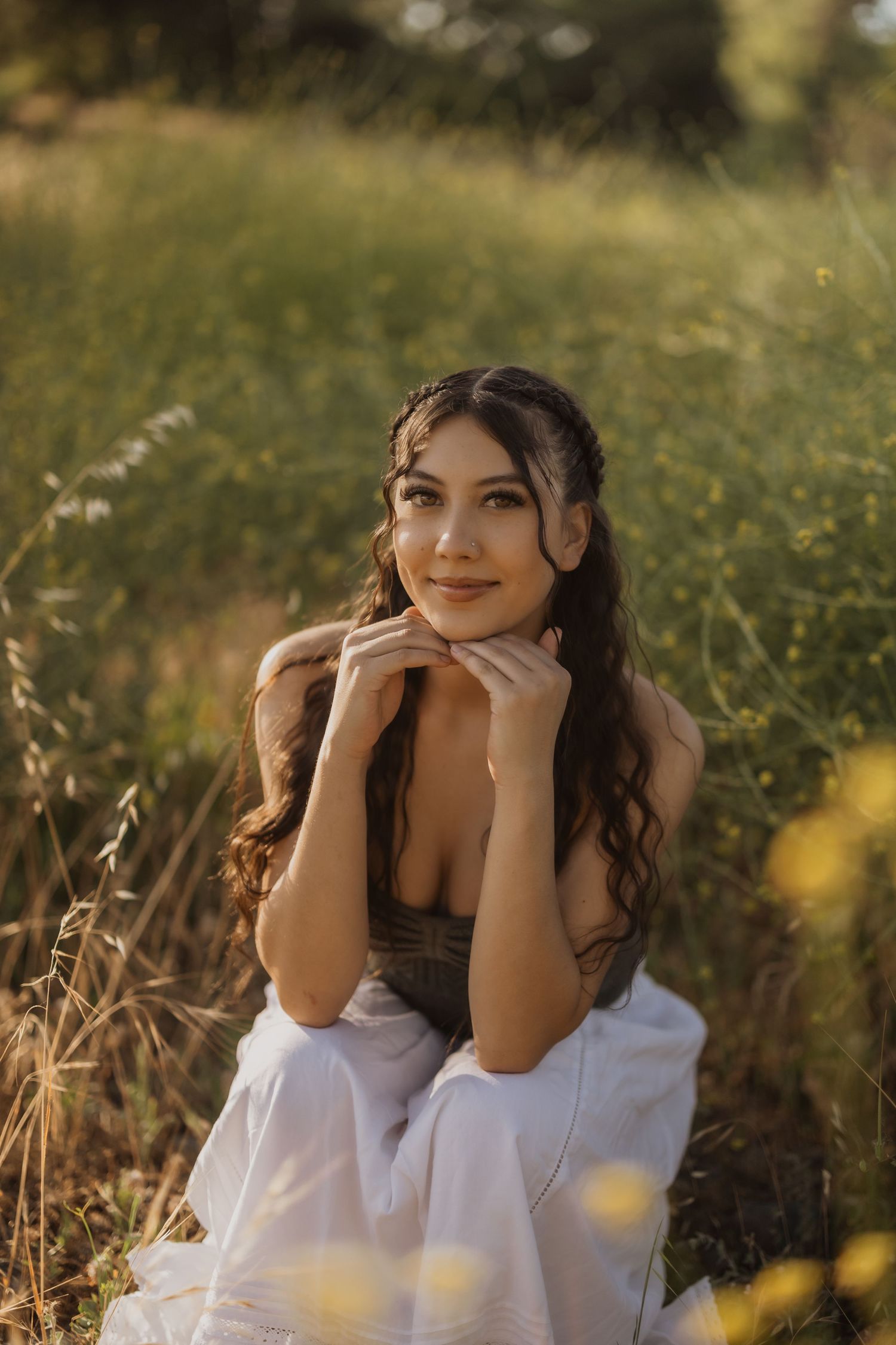 A person in white and brown clothing poses joyfully in a sunlit meadow with wildflowers and tall grass.
