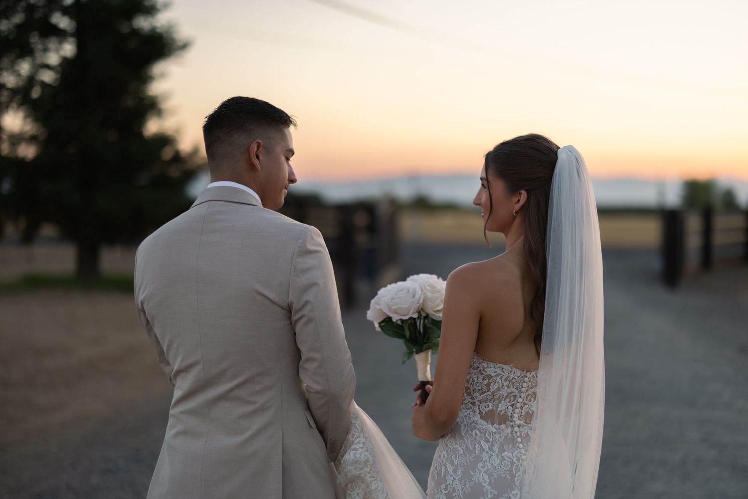 A couple in wedding attire stands together at sunset on a rural path.