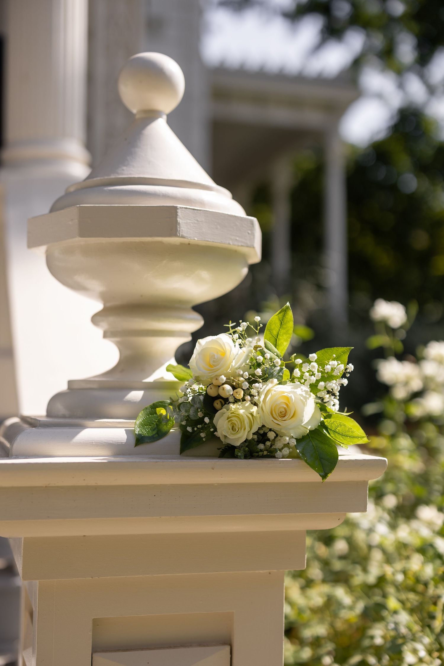 White roses and floral arrangements adorn an elegant white porch railing in sunlight.