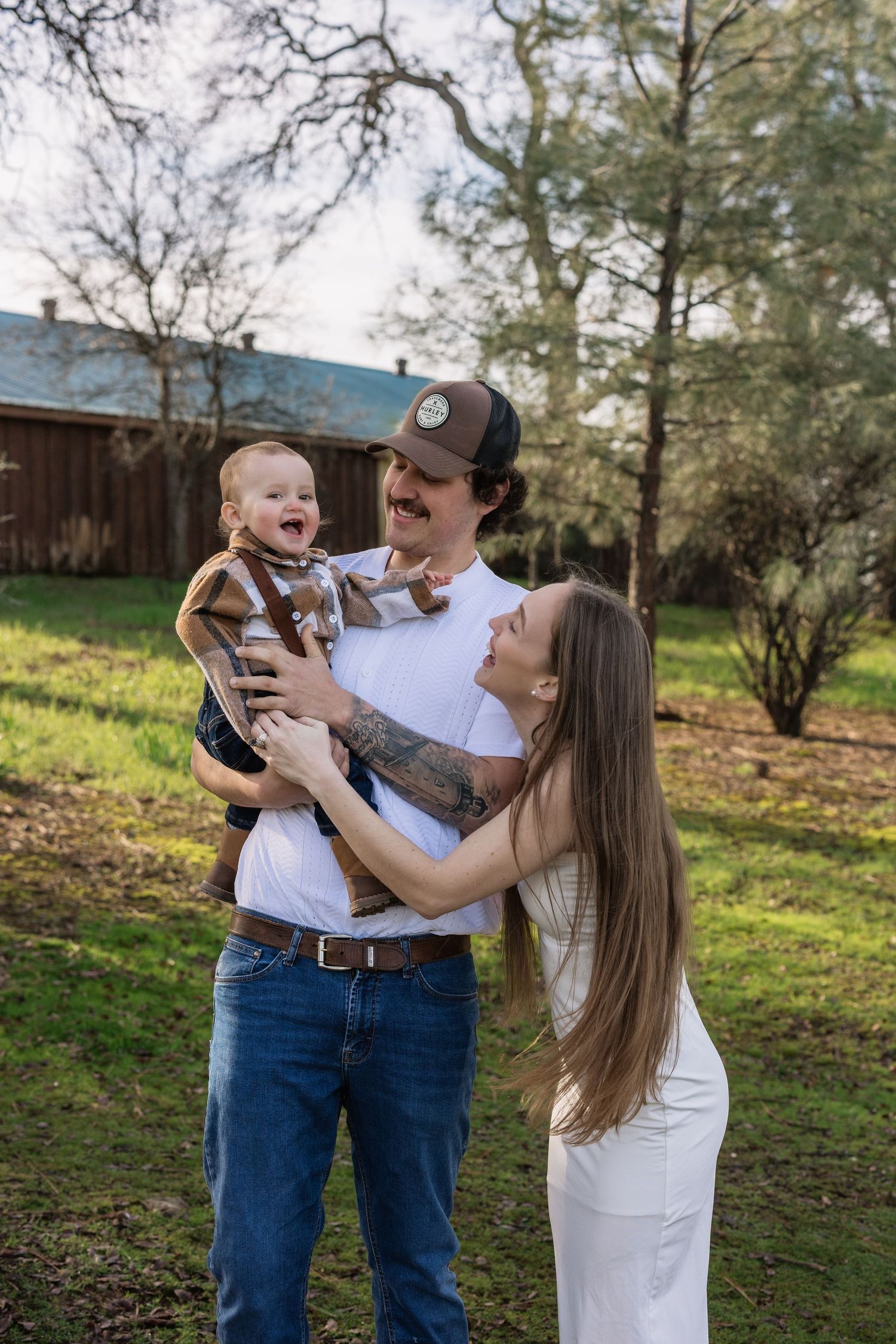 Happy young family with tattooed father in cap holds laughing baby while mother smiles outdoors.