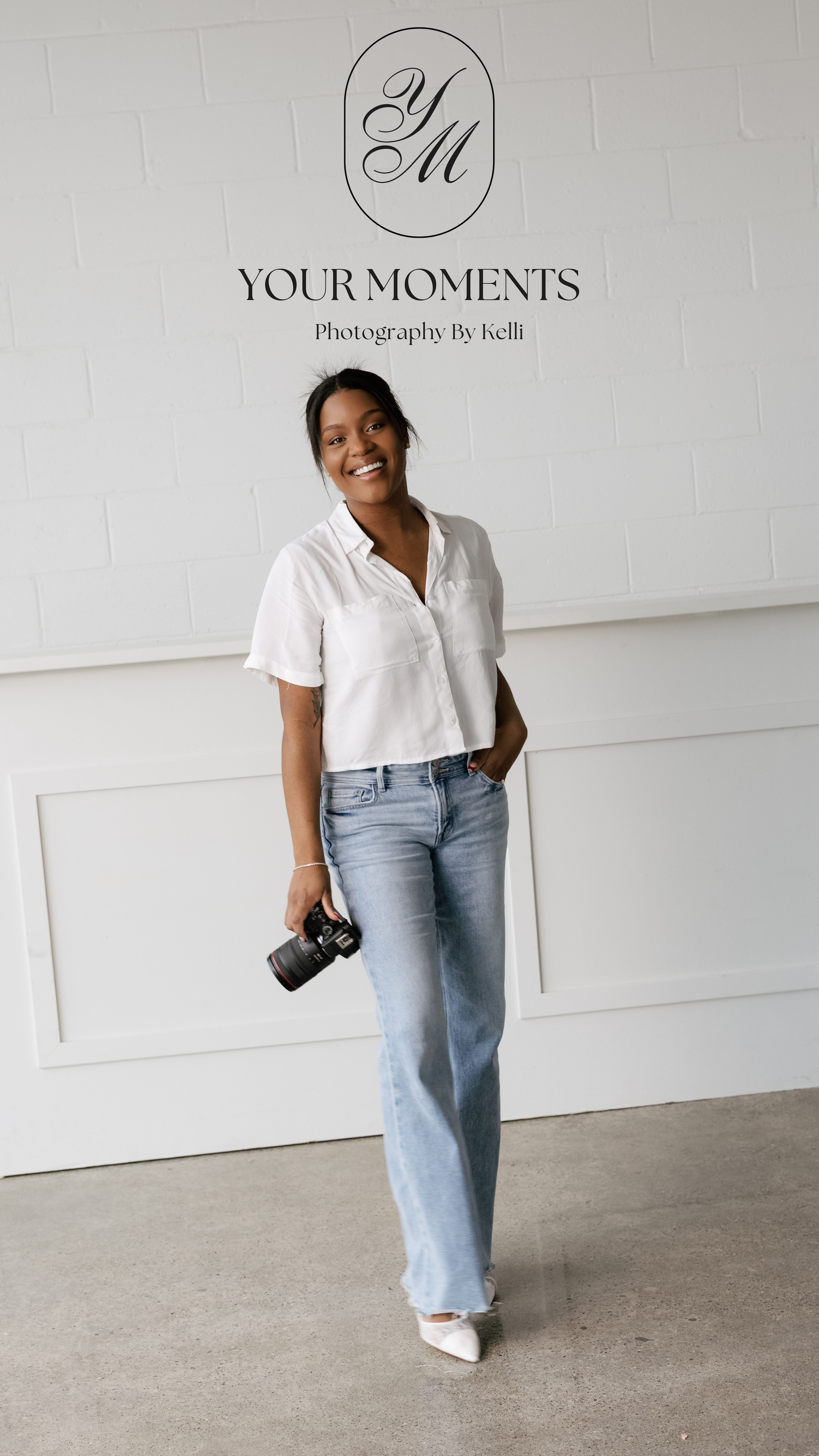 Professional photographer in casual white top and jeans poses against white wall with camera in hand.