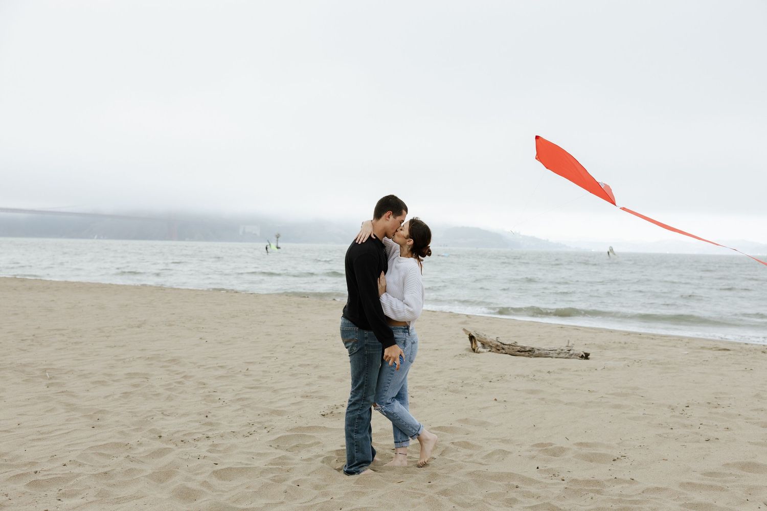 Couple kissing on a sandy beach with a red kite flying nearby on a cloudy day.