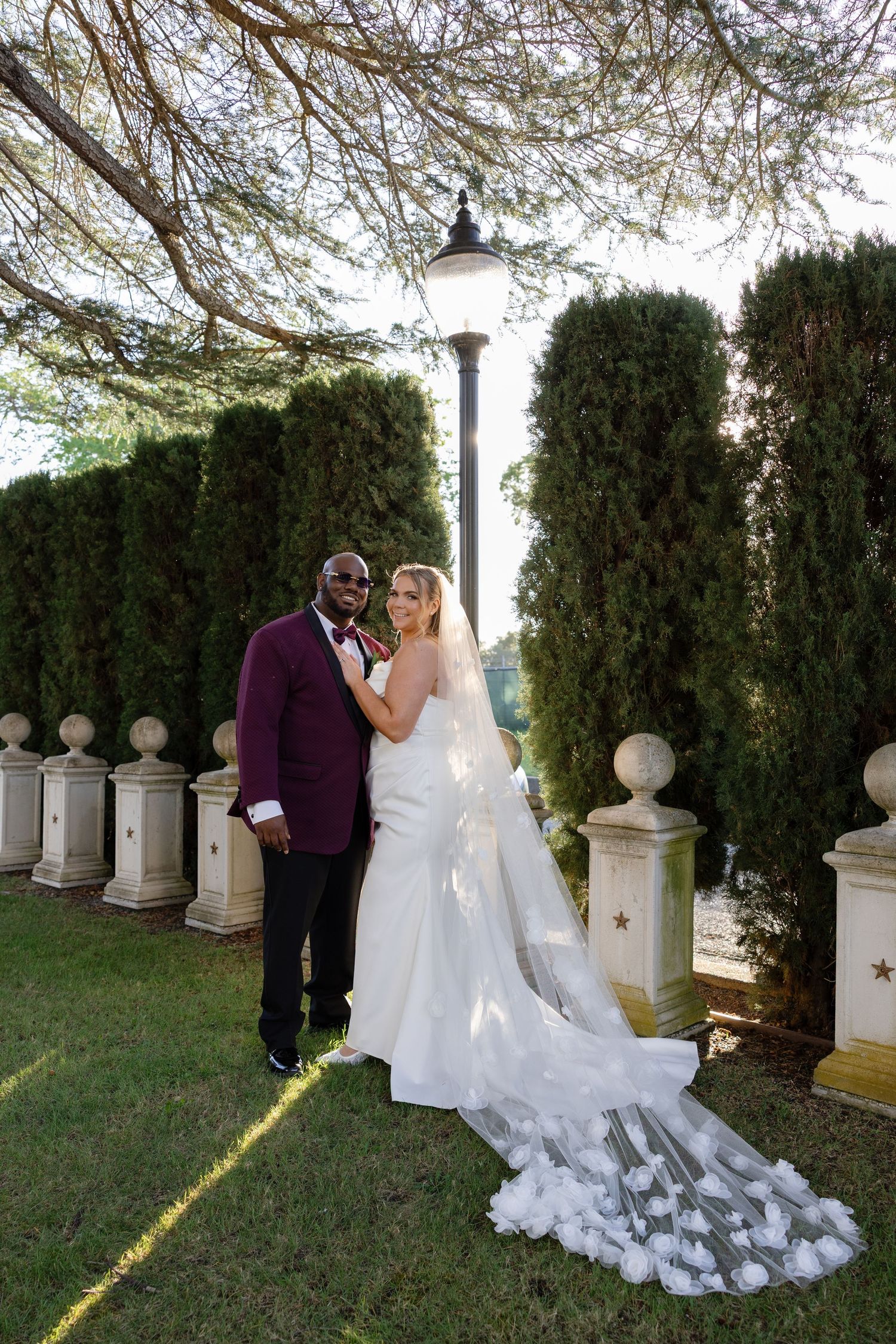 Bride and groom pose elegantly in a garden wedding venue with tall cypress trees and a vintage lamp post.