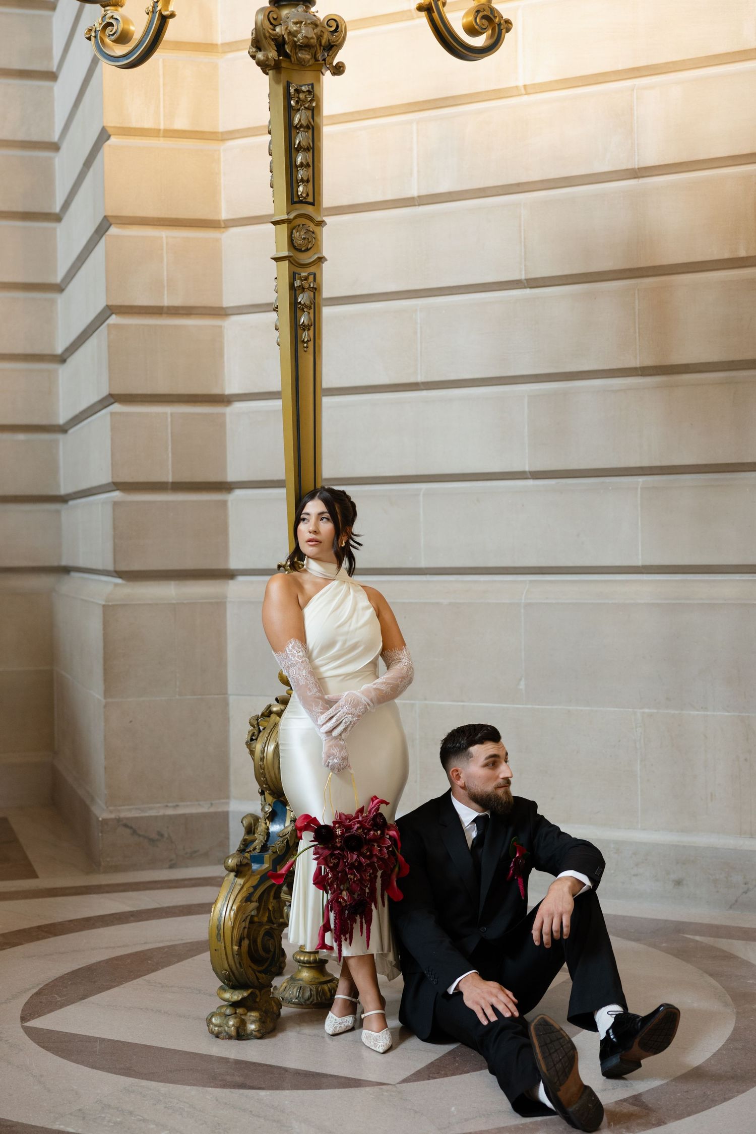 Bride in white halter gown with lace gloves and groom in black suit pose by ornate gold pillar in elegant venue.