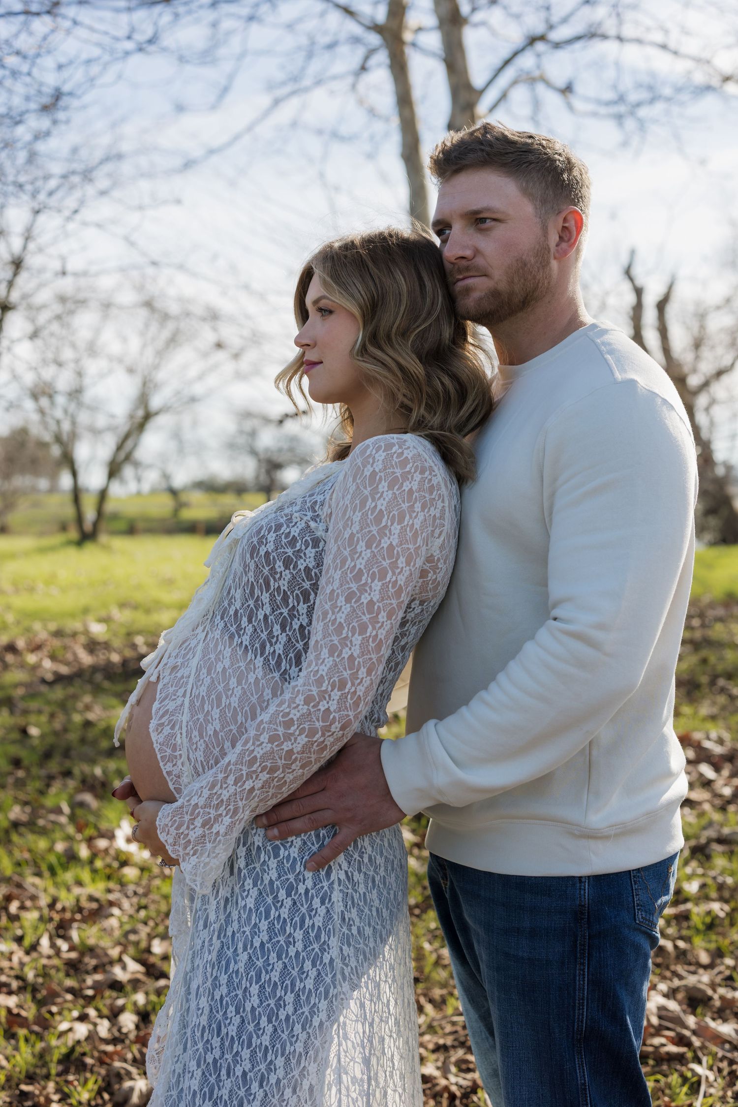 A couple embraces outdoors in a white lace dress and sweater against a sunlit natural backdrop.