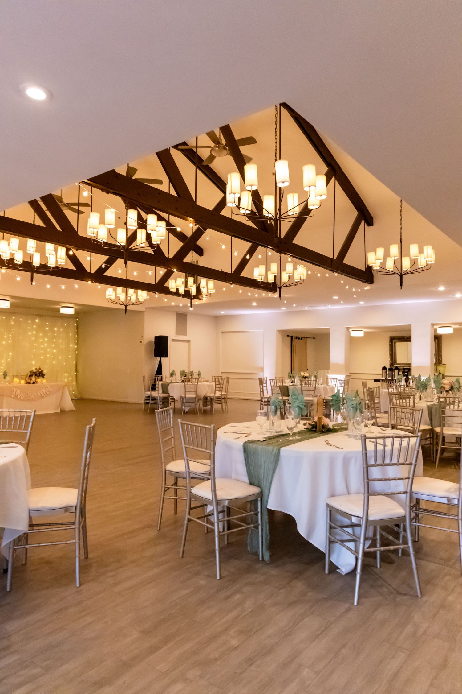 Modern wedding reception venue with exposed beam ceiling and elegant chandelier lighting over round tables.