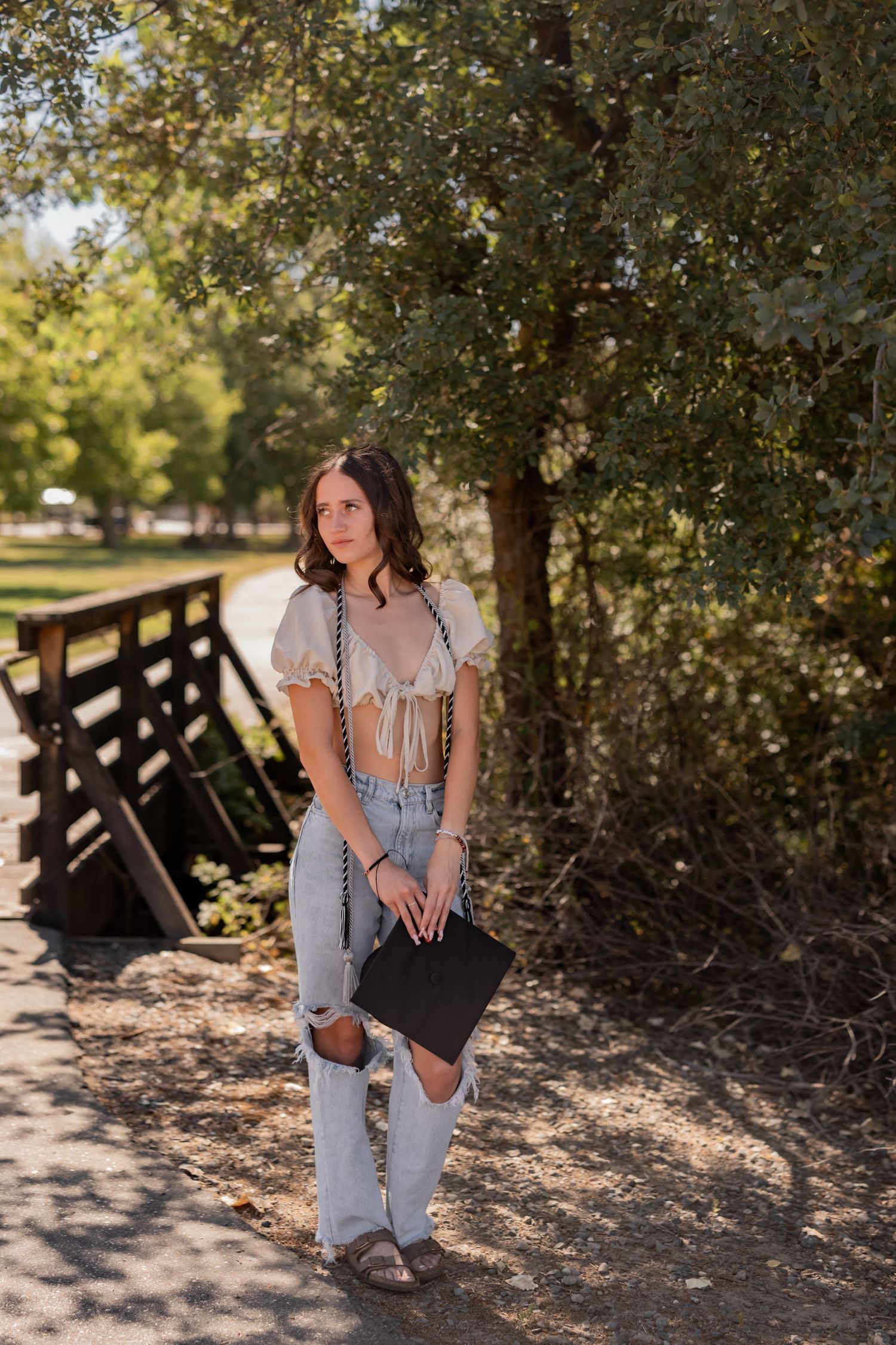 Graduate in casual attire holds diploma while posing on wooden bridge pathway.