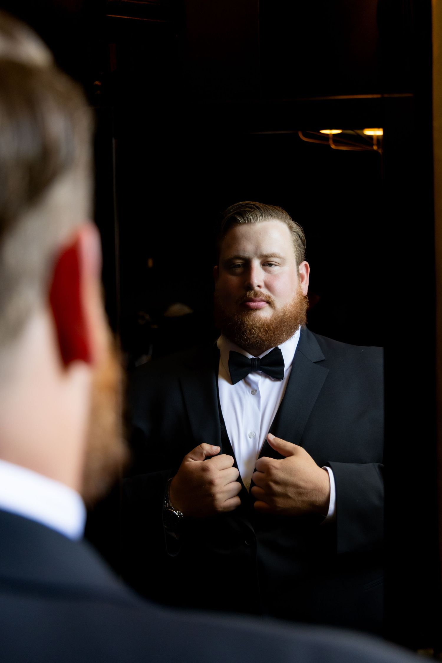 Reflection of a groom adjusting his bowtie in a dark-toned mirror.