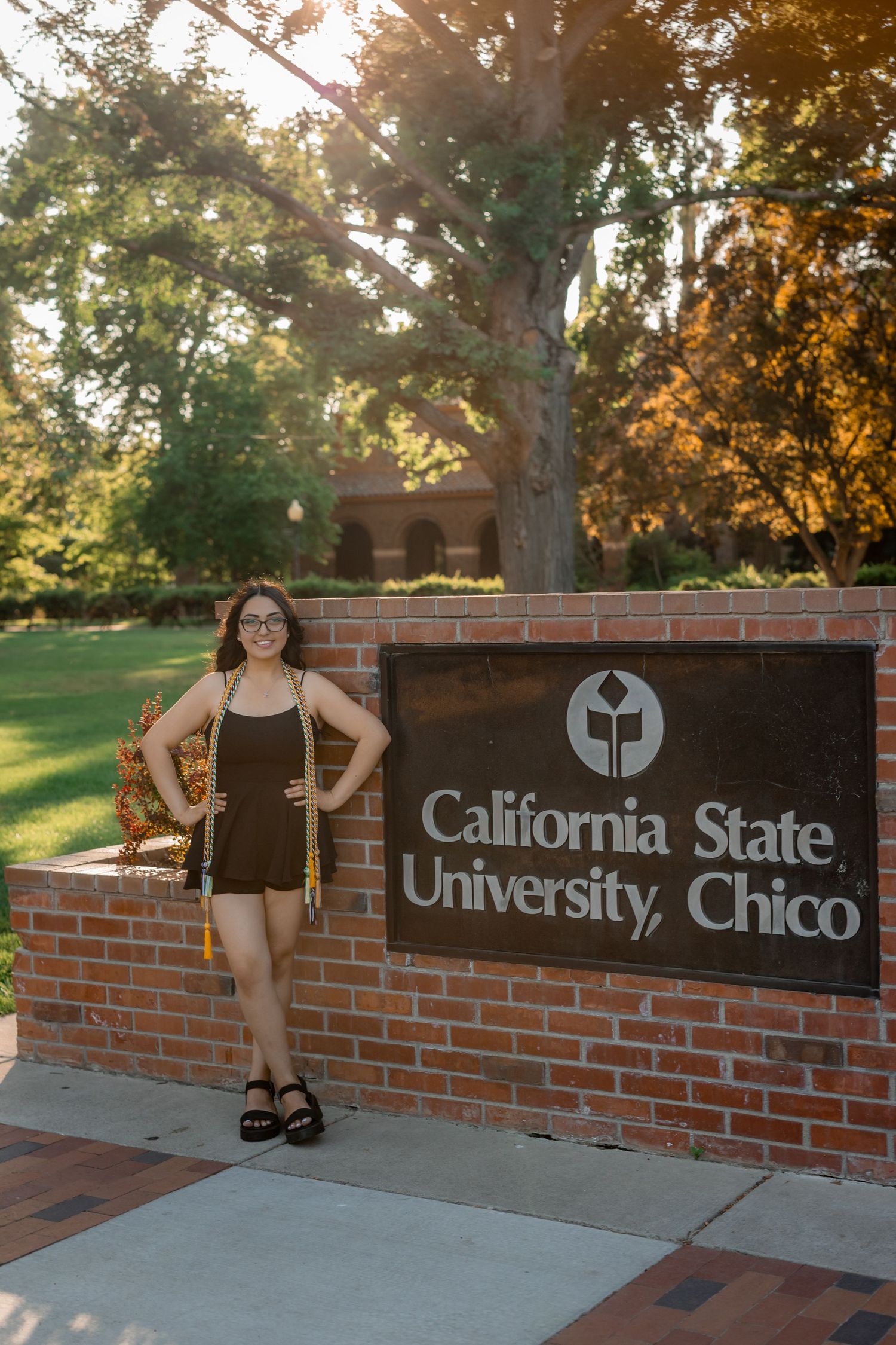 Graduate poses at California State University Chico brick entrance sign in autumn setting.