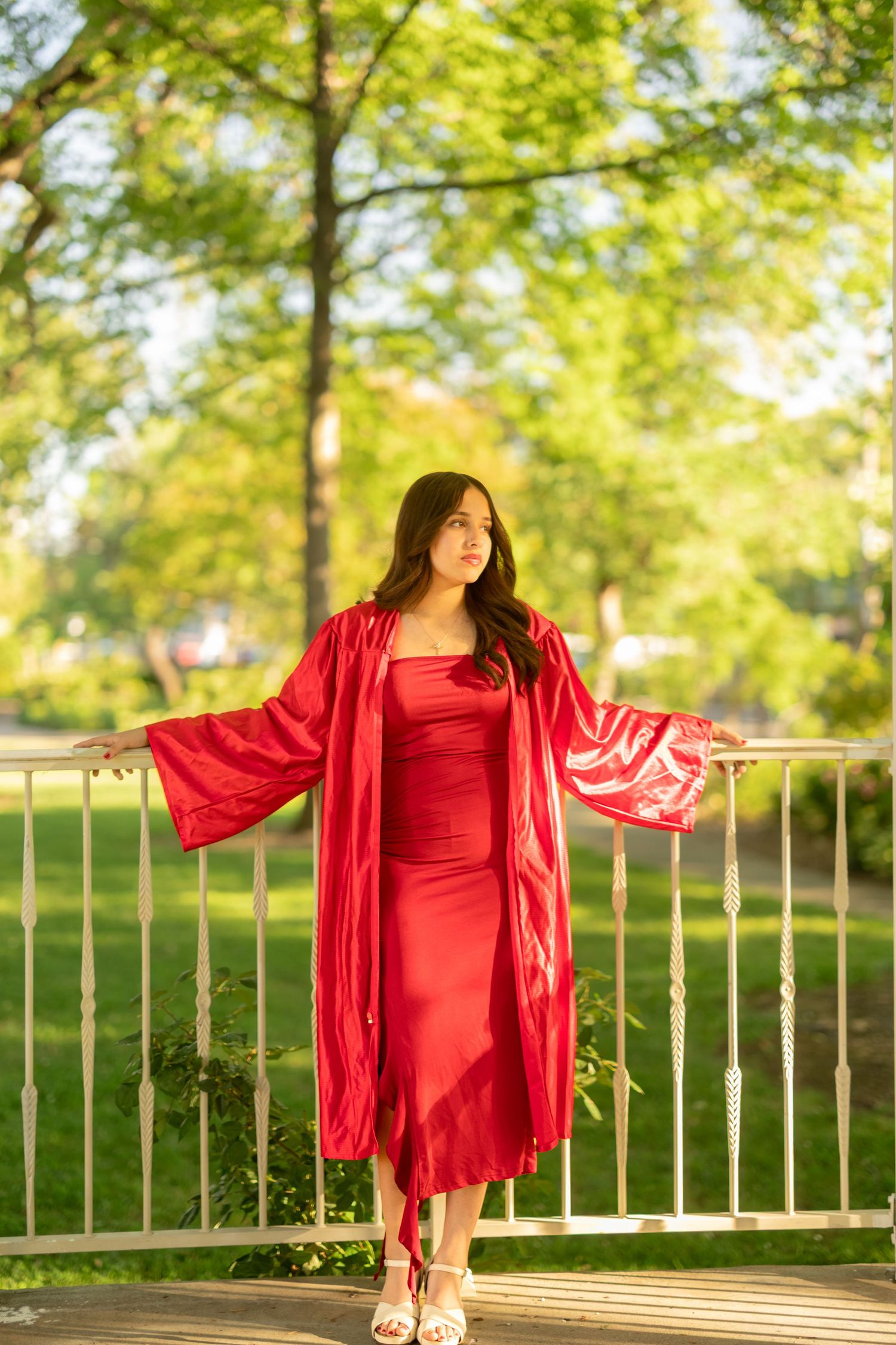 Graduate poses in flowing red graduation gown against green park backdrop.