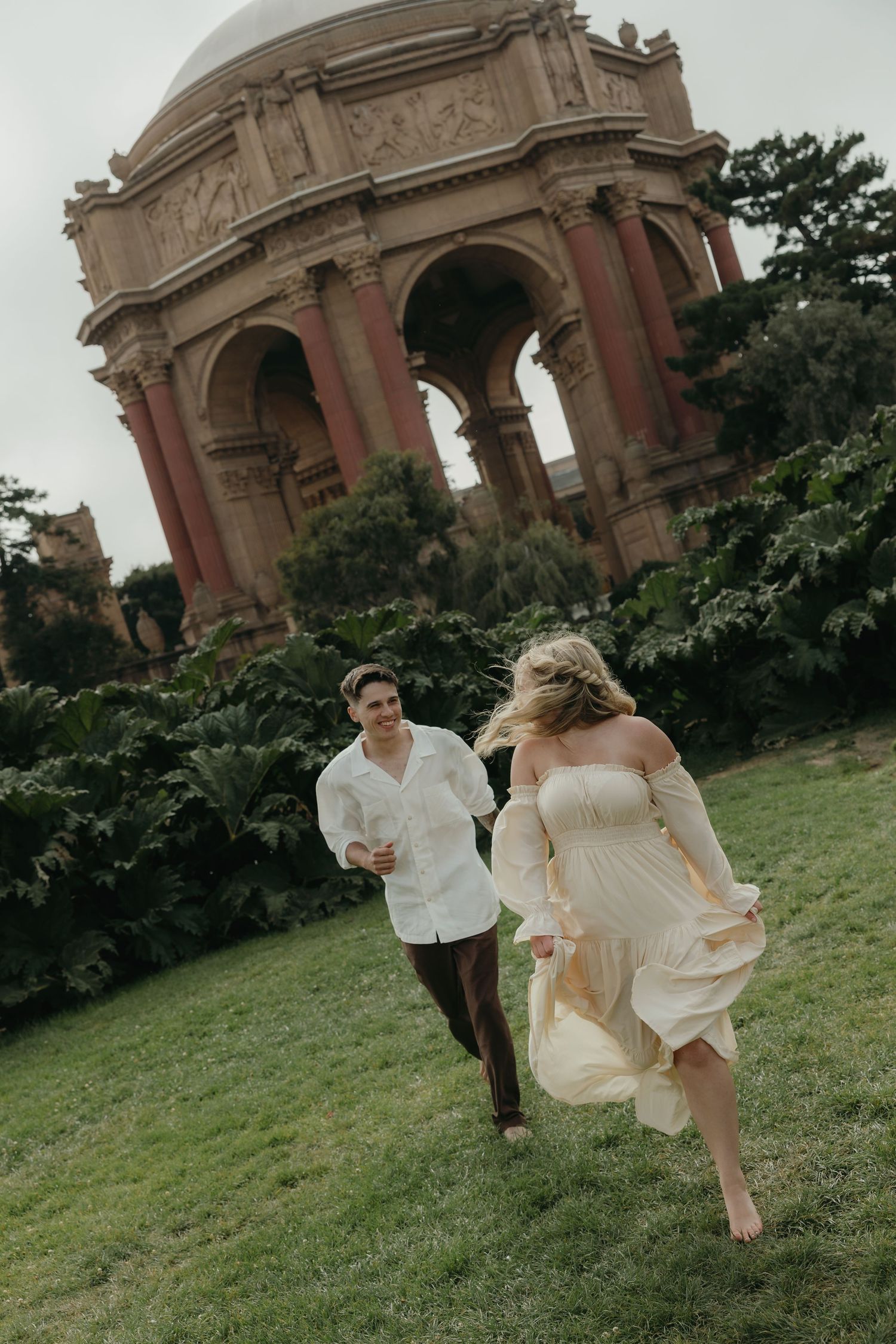 Couple running on grass near the Palace of Fine Arts rotunda, woman in flowing cream dress, man in white shirt.