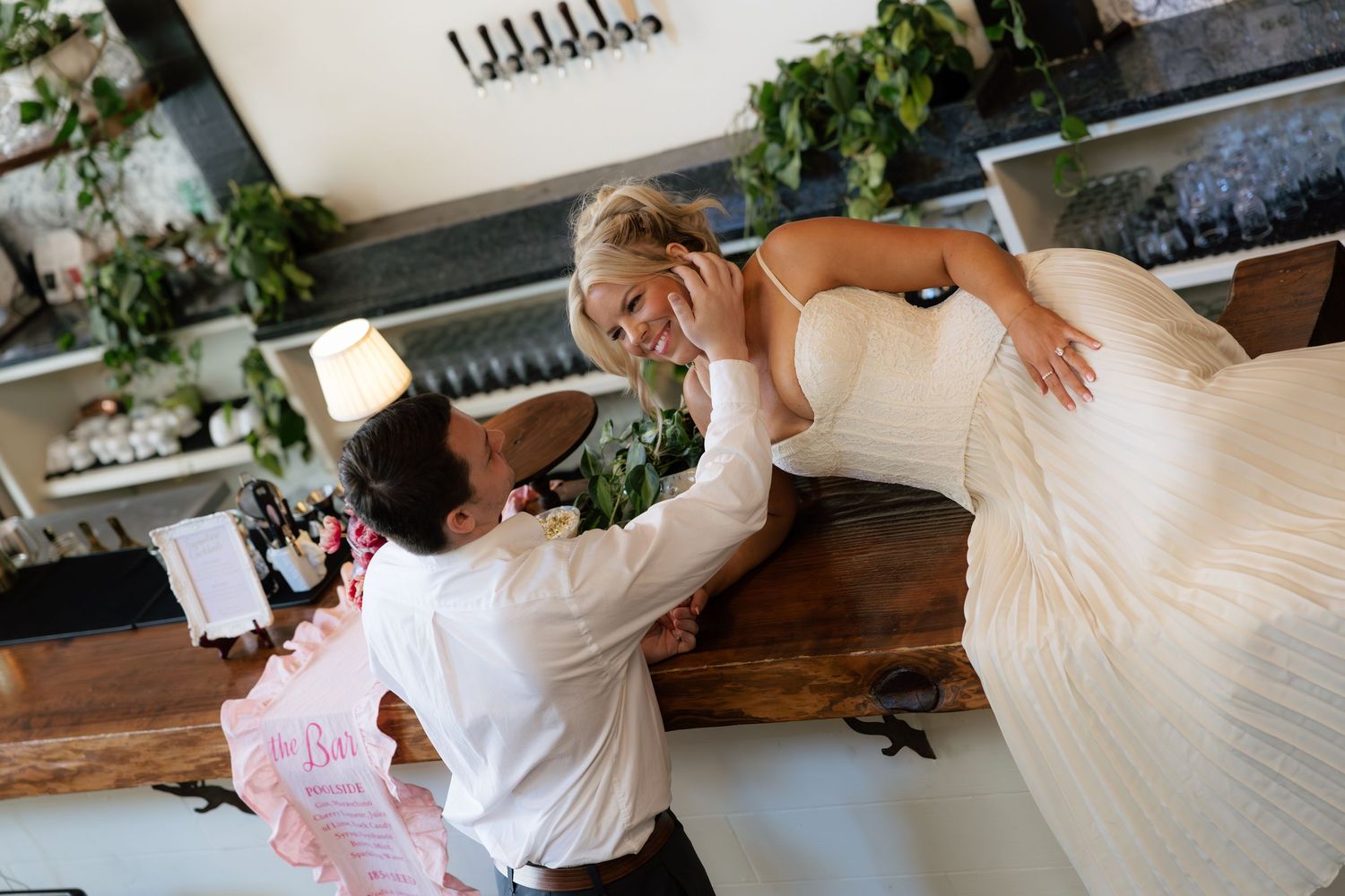A groom tenderly touches his bride's face as she leans on a wooden bar in an elegant wedding venue.