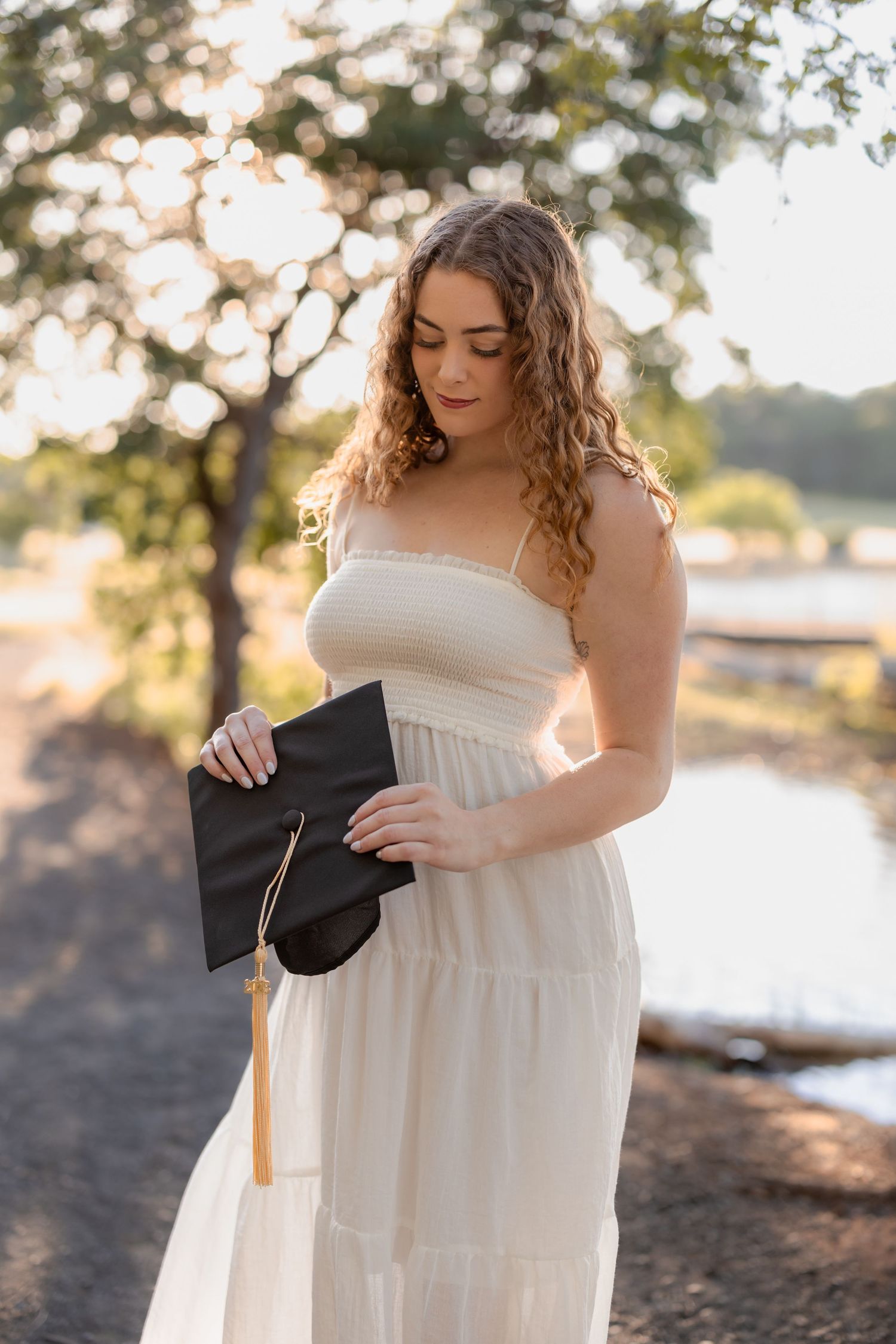 Graduate in white dress holds diploma while posing at sunset by a lake.
