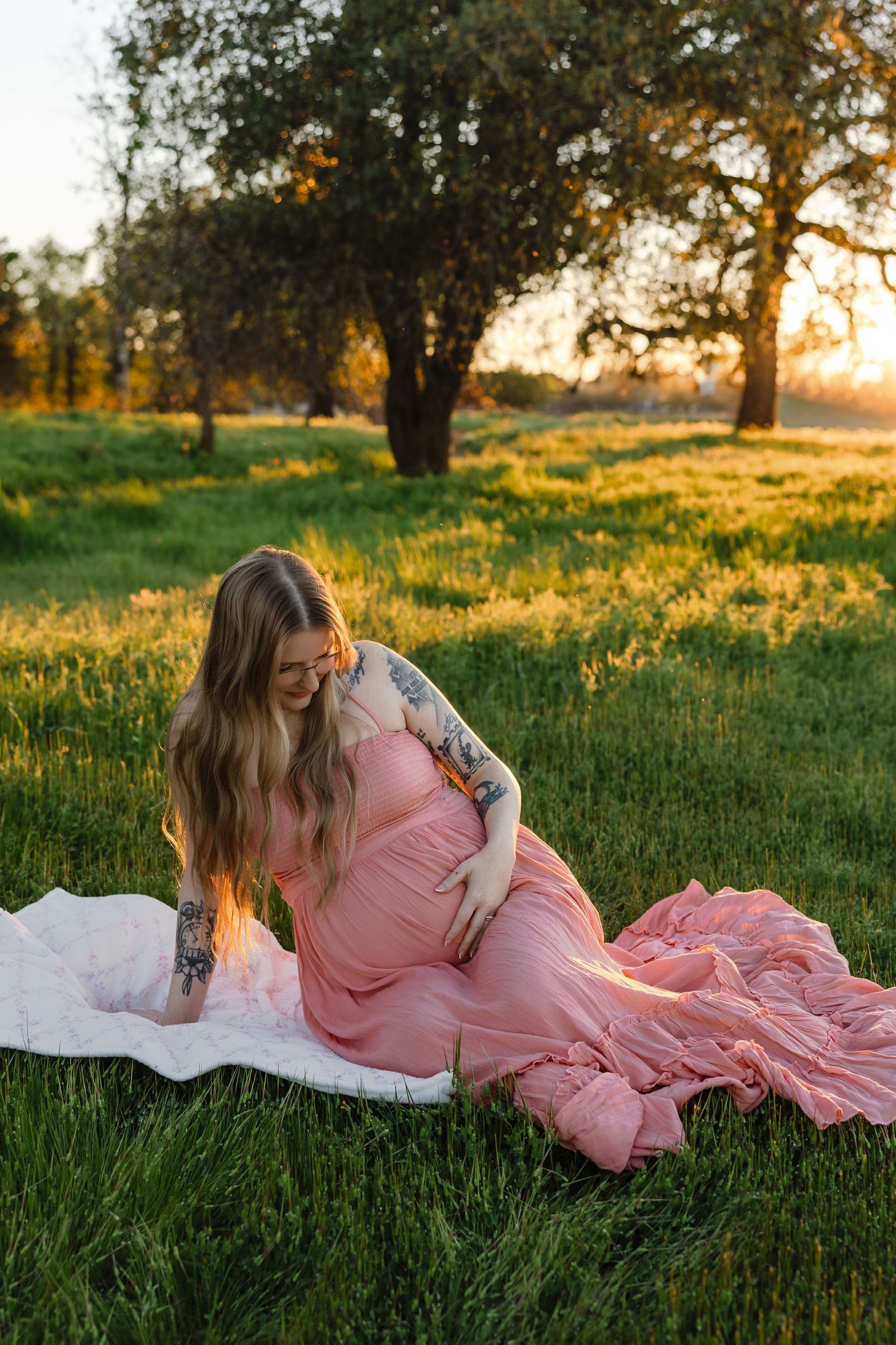 Pregnant woman in pink dress sitting on grass at golden hour sunset in a lush green field with trees.