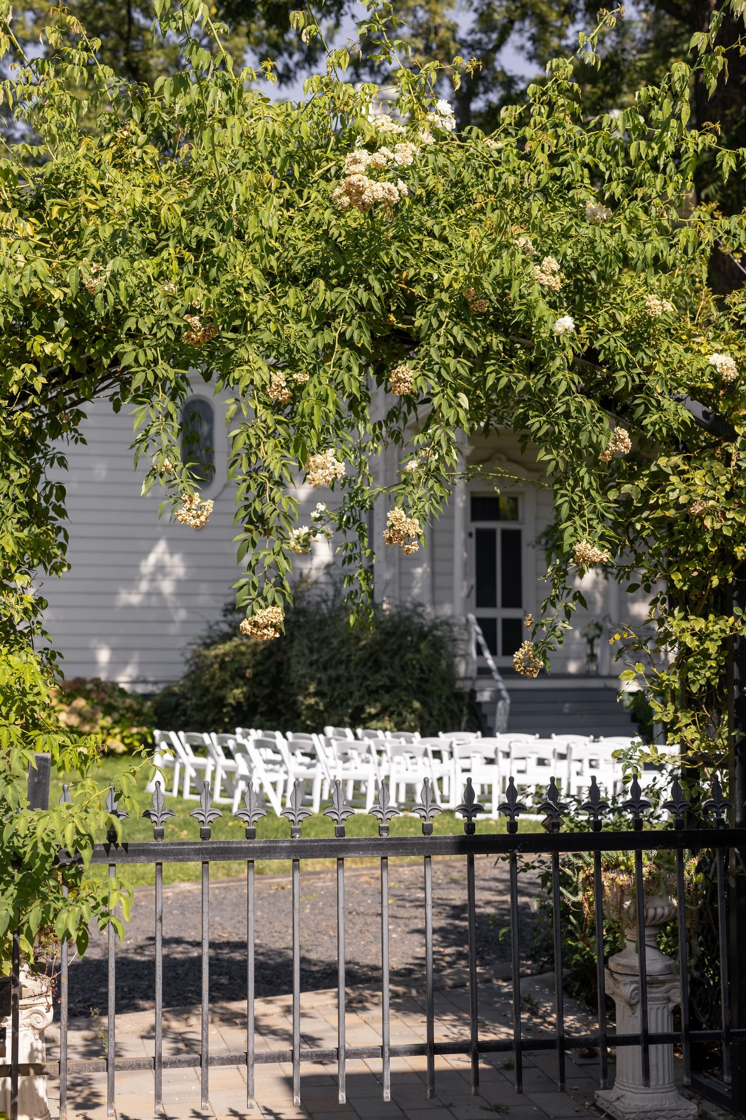 White ceremony chairs arranged outdoors at a Victorian mansion under shady trees.