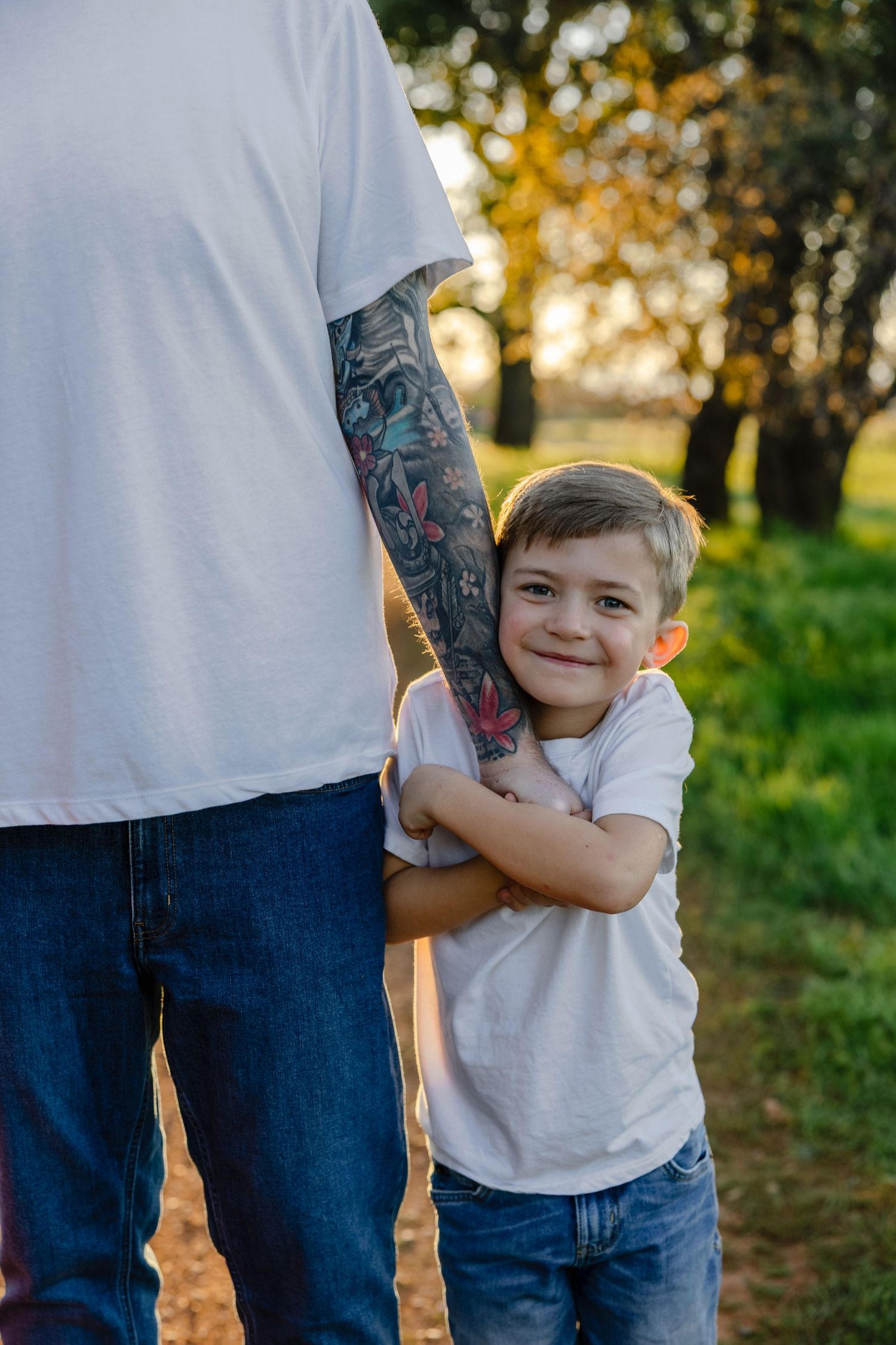 Smiling young boy hugging tattooed arm of adult in white t-shirt and jeans outdoors at golden hour.